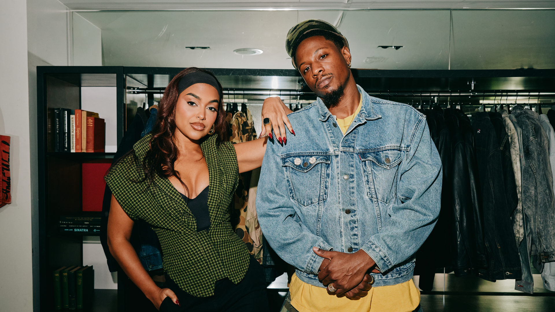 A woman in a green top and a man in a denim jacket stand in a stylish room, with clothing racks and books in the background.