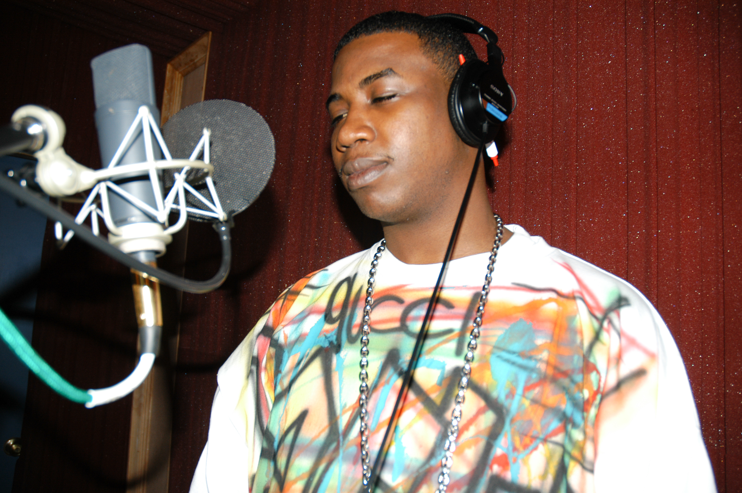 Gucci Mane wearing headphones and a colorful shirt stands in front of a microphone in a recording studio.