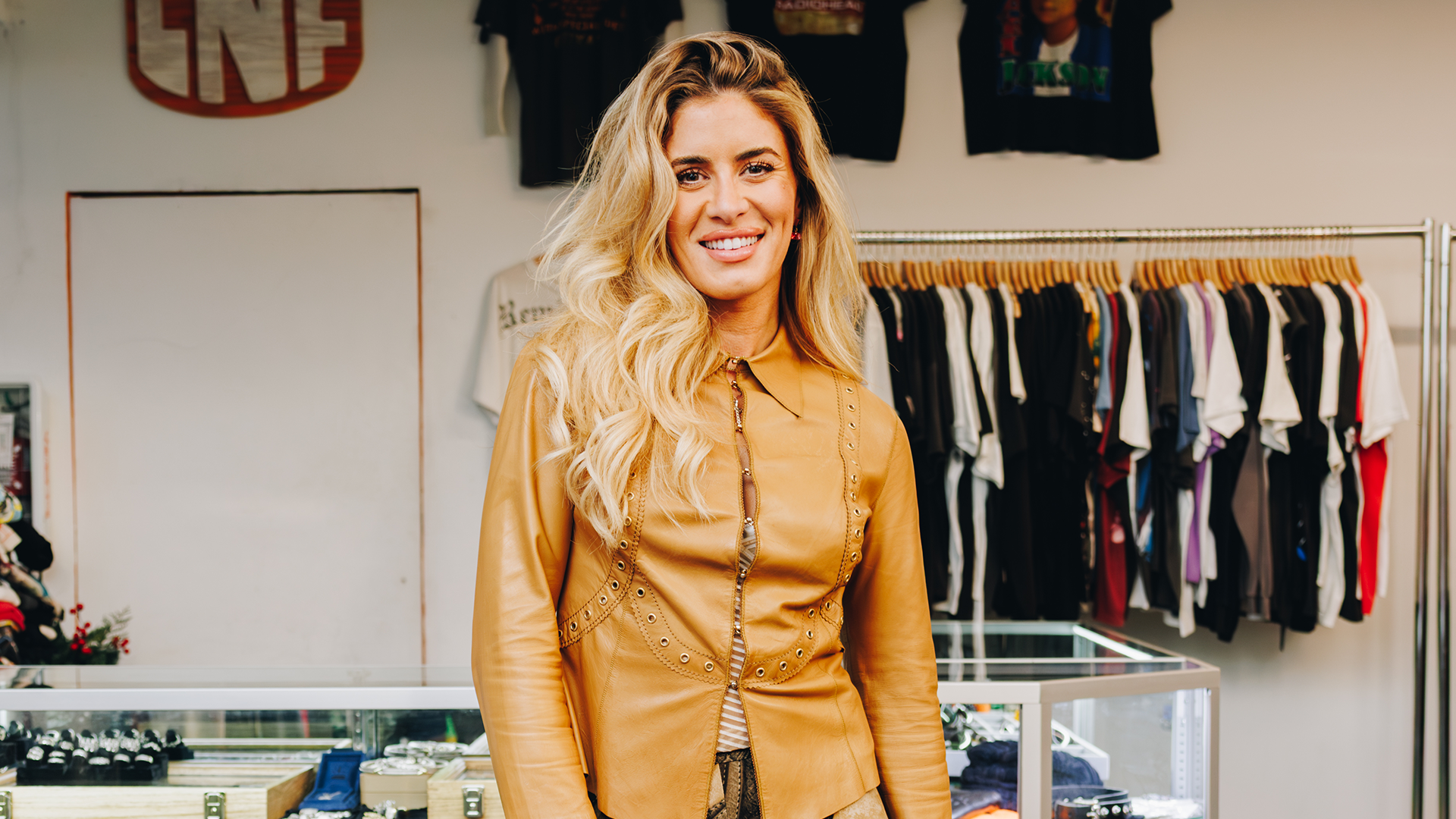 A woman with long blonde hair in a tan leather jacket stands in a clothing store with racks of shirts in the background.