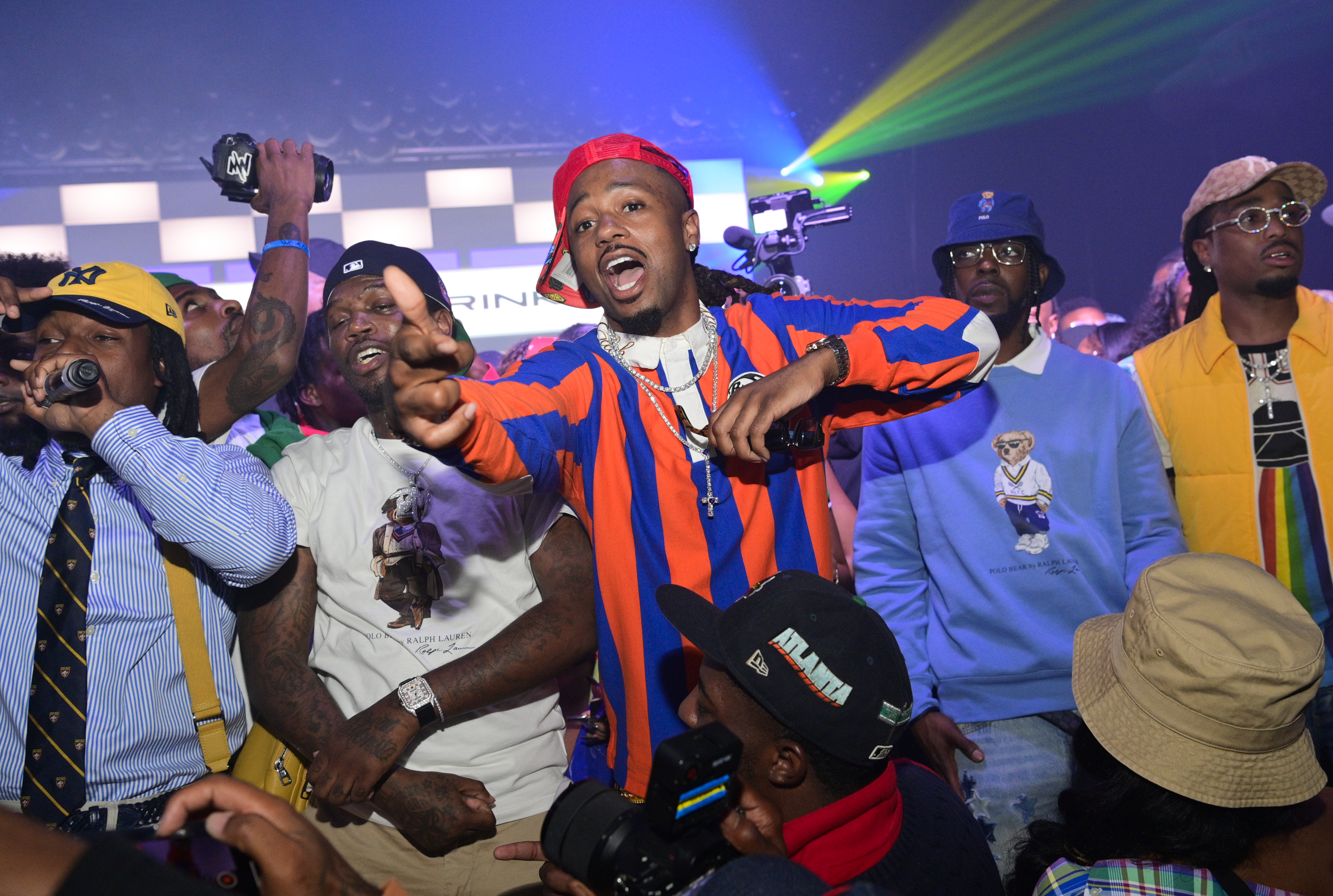 Metro Boomin at a party wearing a red cap and striped shirt energetically pointing, surrounded by others in colorful attire.