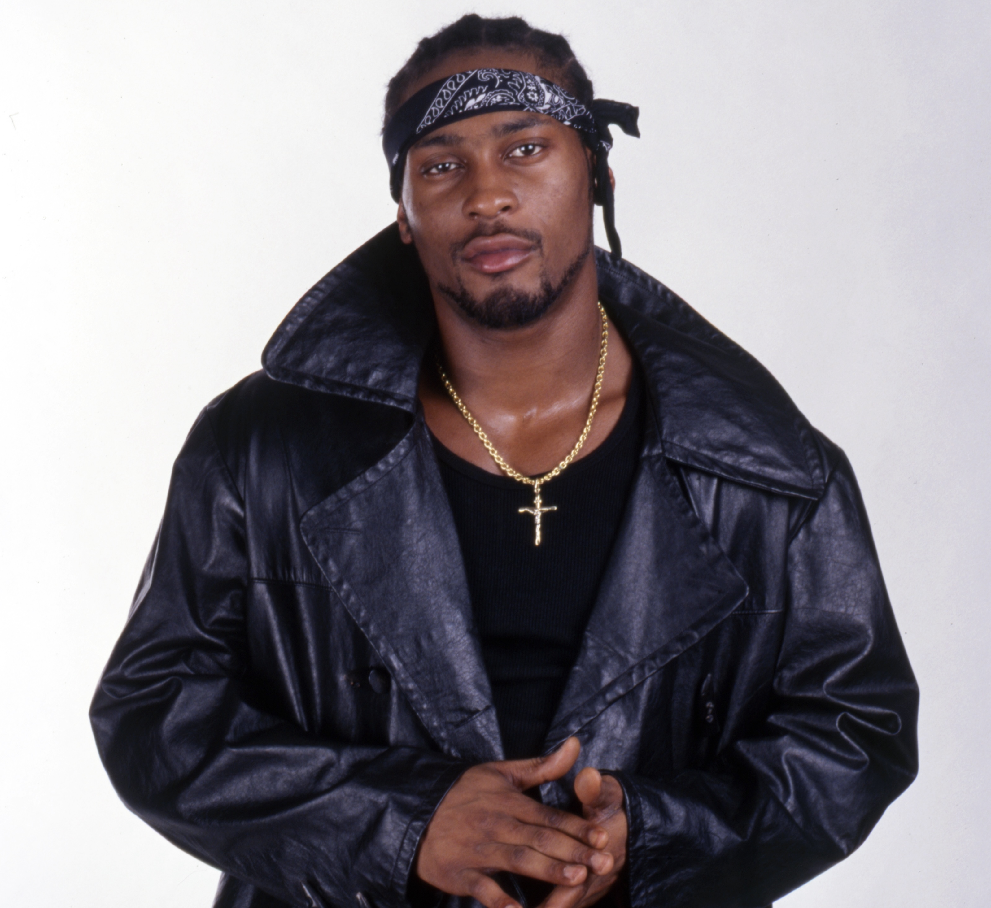 A man wearing a black leather jacket, black shirt, and a bandana, with a gold cross necklace, standing against a plain background.