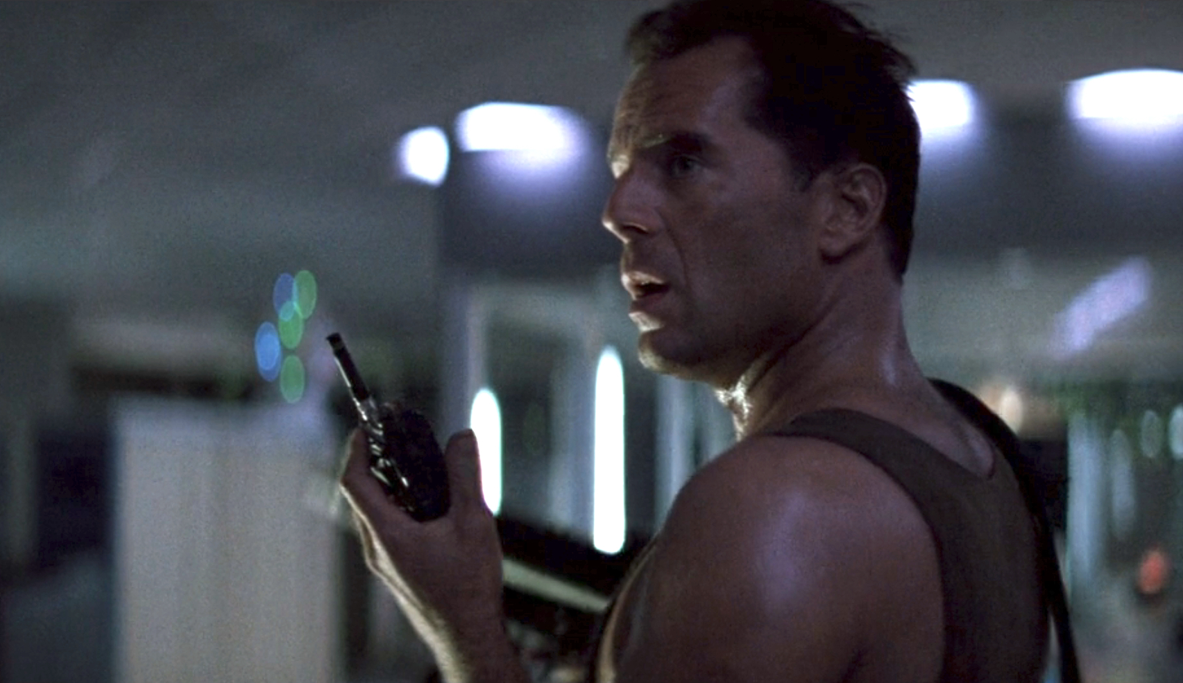 Bruce Willis in a scene from "Die Hard," holding a walkie-talkie and wearing a tank top, with a tense expression.