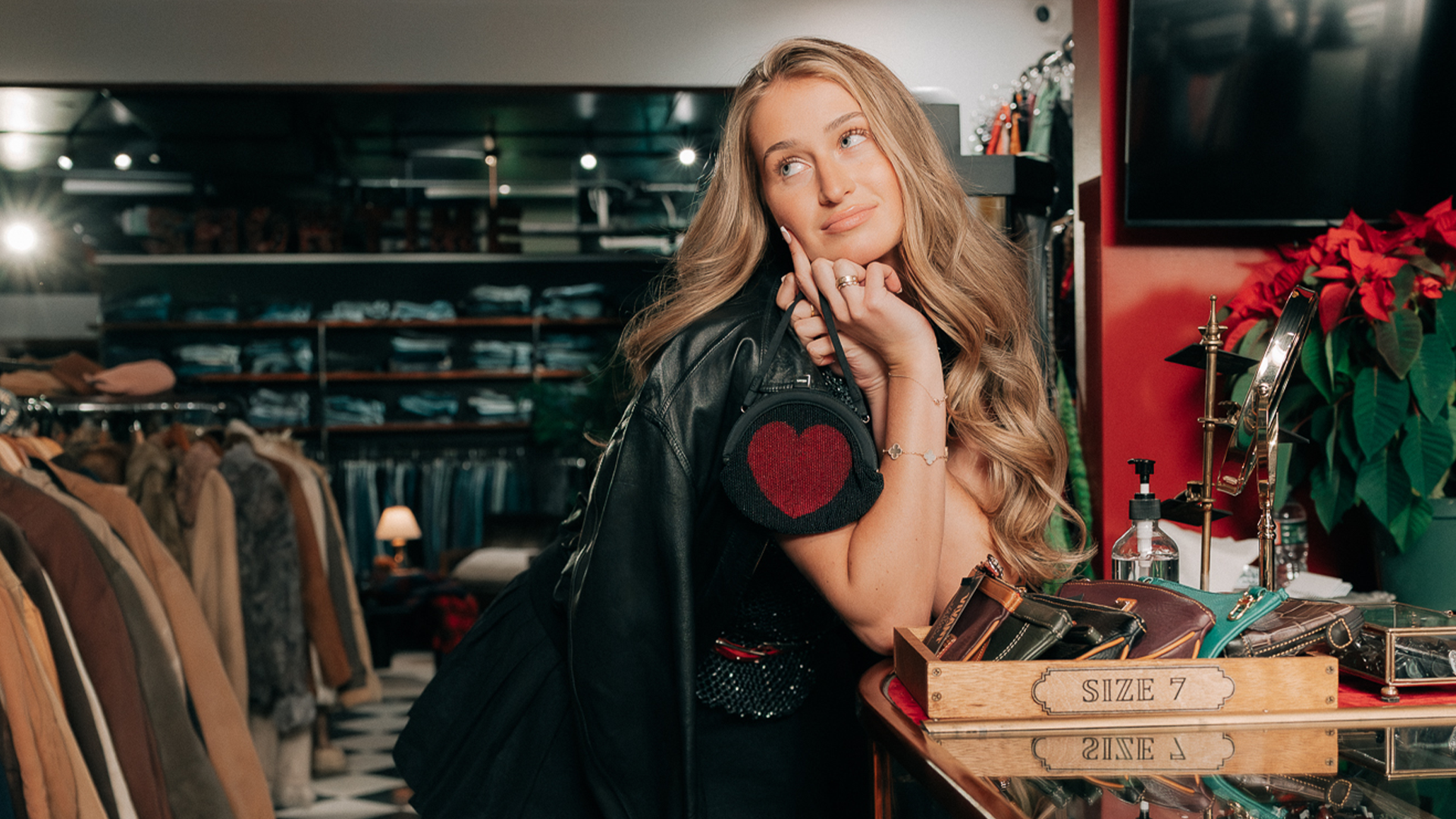 A woman with long blonde hair in a store, leaning on a counter, holding a small bag with a heart design. Shelves and clothing in the background.