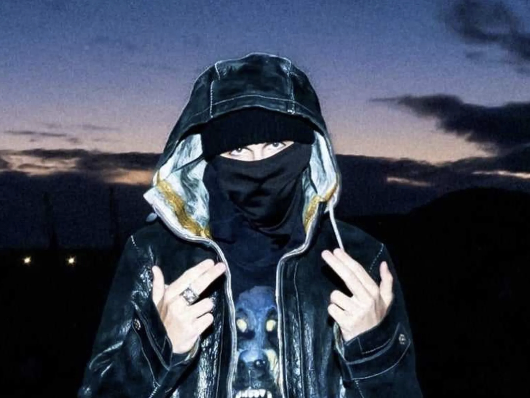 EsDeeKid in a hooded jacket and mask stands against a twilight sky, pointing with both hands.