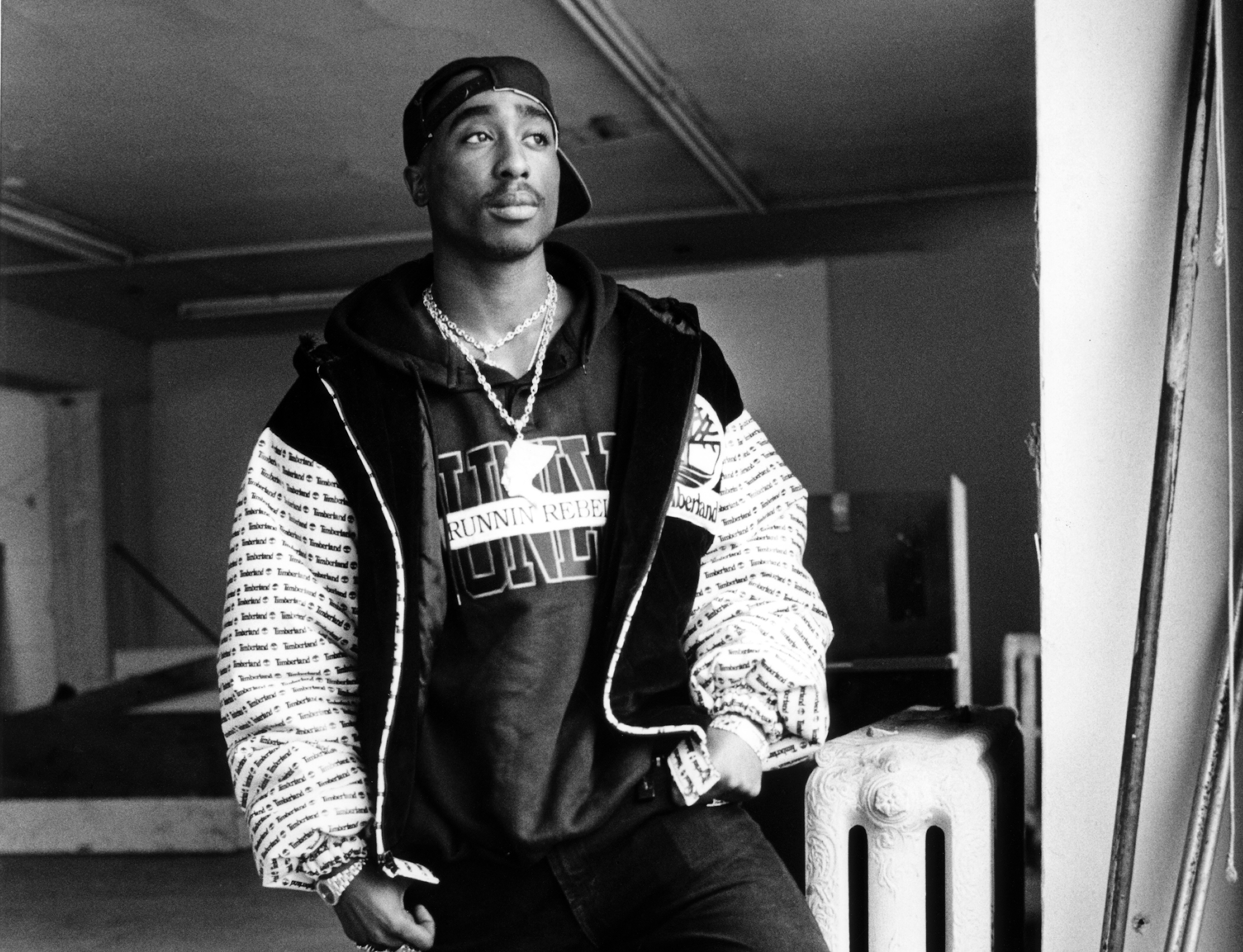 Black and white photo of Tupac Shakur wearing a cap backward, a hoodie, and a jacket, standing in a room with a thoughtful expression.