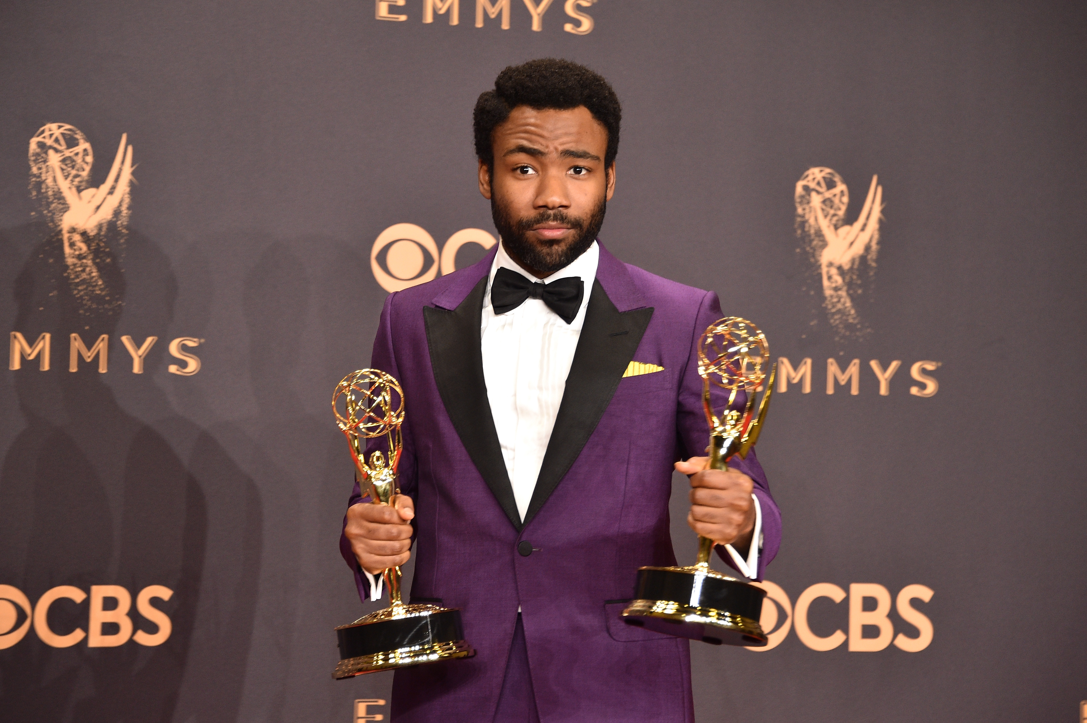 Donald Glover holding two Emmy awards, wearing a purple tuxedo with a bow tie, at the Emmy Awards event.