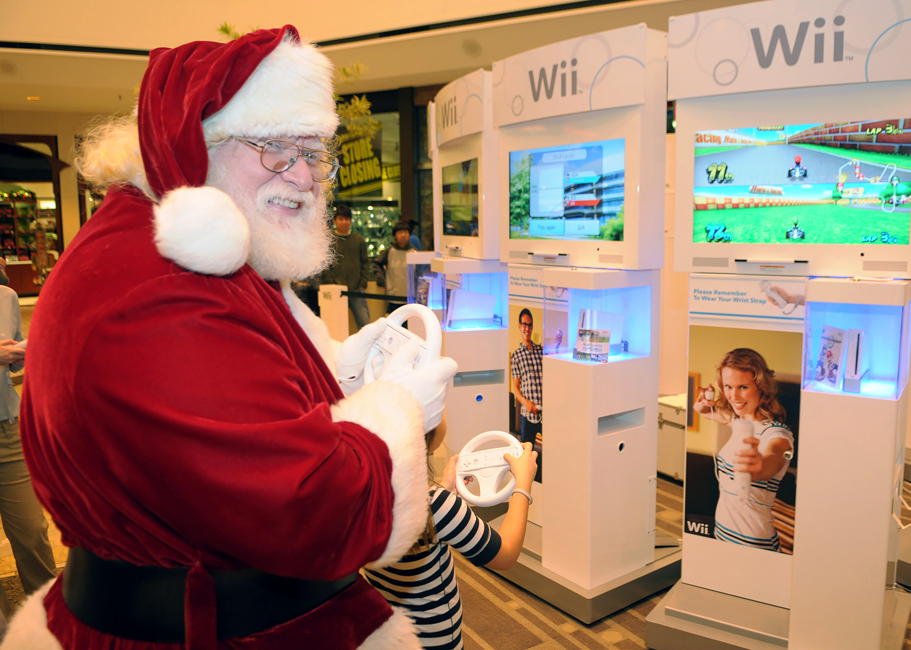 Santa Claus playing a Wii game at a store display, smiling and holding a controller. A child plays beside him.
