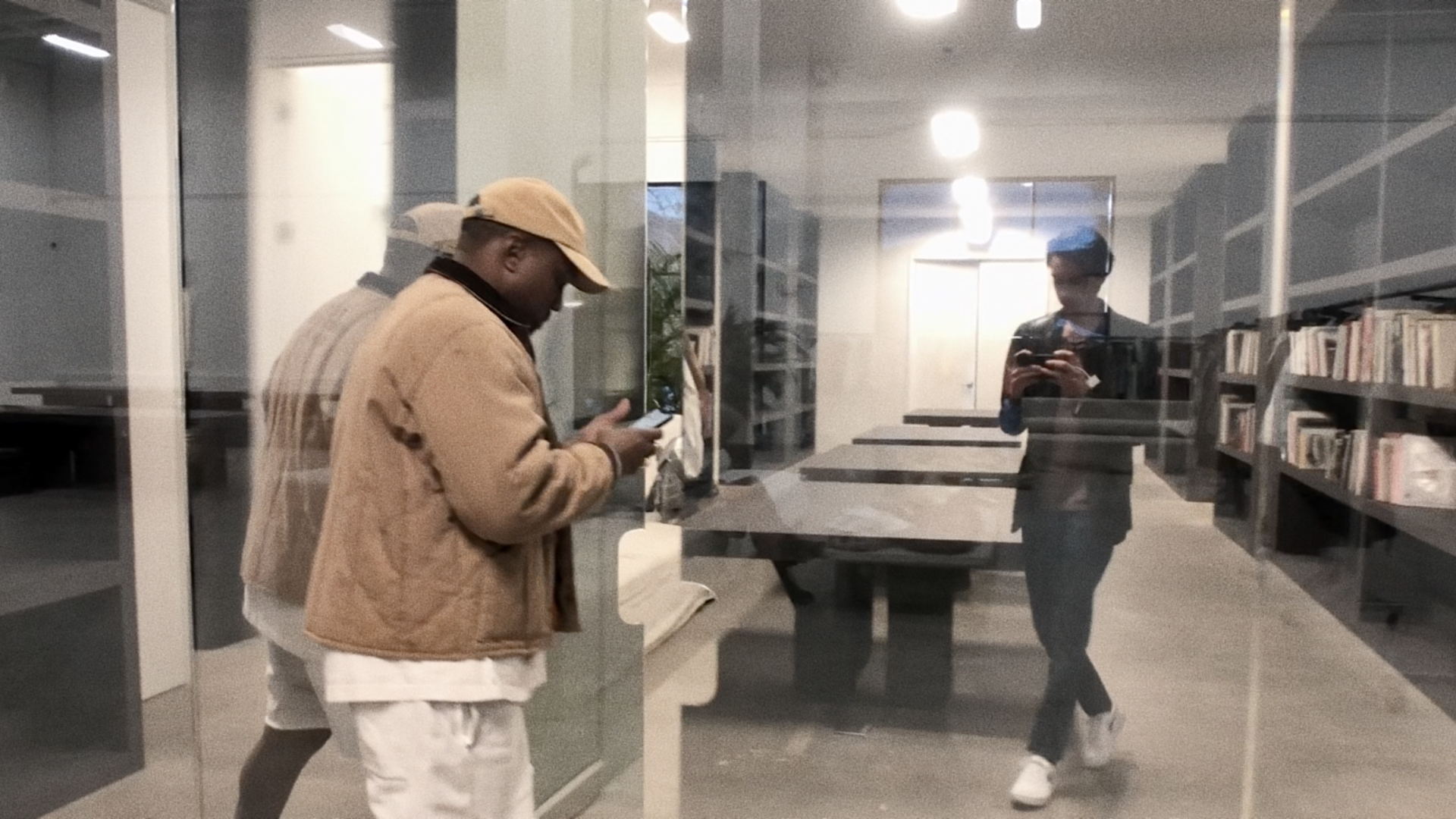 Ye and Nico Ballesteros are seen through a glass wall in a modern library or office. One is using a phone, while the other is reflected in the glass.
