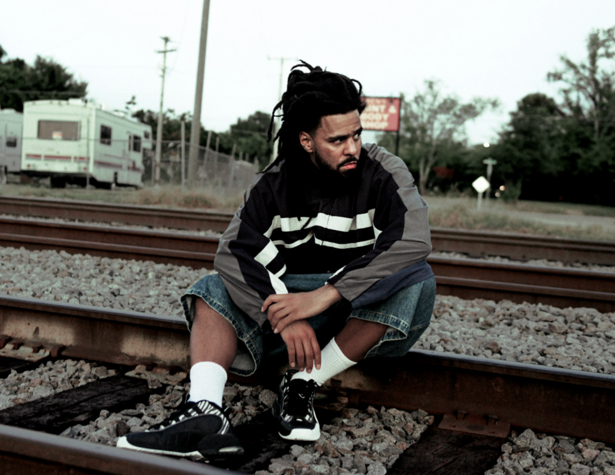 J. Cole, who has dreadlocks, is wearing a striped jacket and denim shorts. He sits pensively on train tracks. His next album is 'The Fall Off' and he's been working on it for about a decade.
