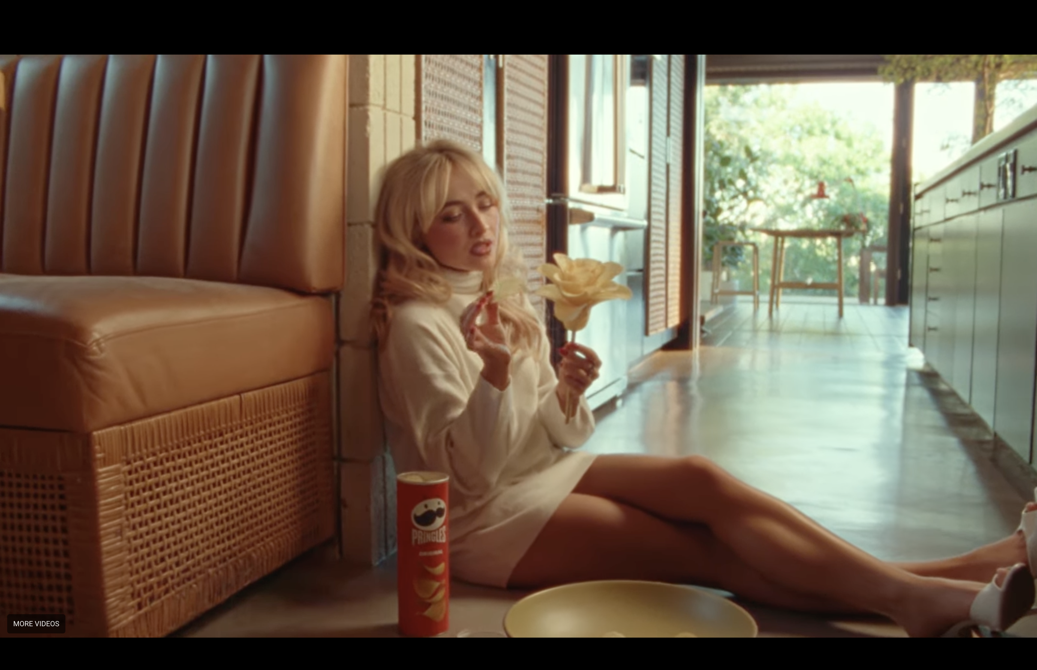 Sabrina Carpenter sits on the floor in a cozy room, holding a flower. A can of Pringles is nearby. Warm lighting fills the space.