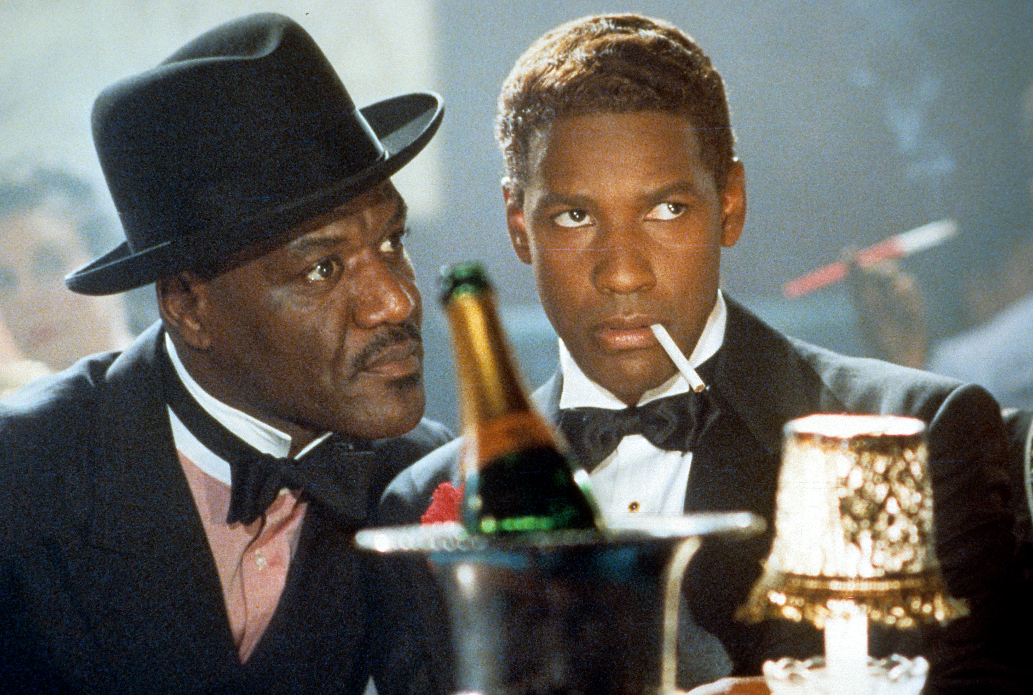 A scene from a film with Denzel Washington, dressed in a tuxedo and holding a cigarette, sitting beside another man in a suit and hat.