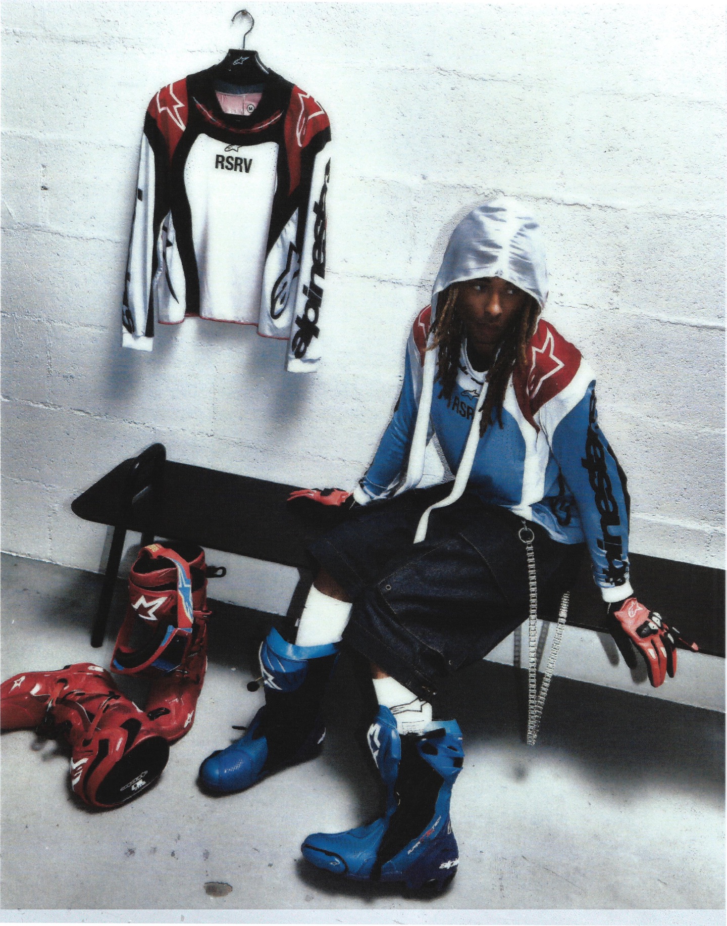 Person in motocross gear with a hoodie and boots sits on a bench. A jersey hangs on the wall, and red boots are on the floor.