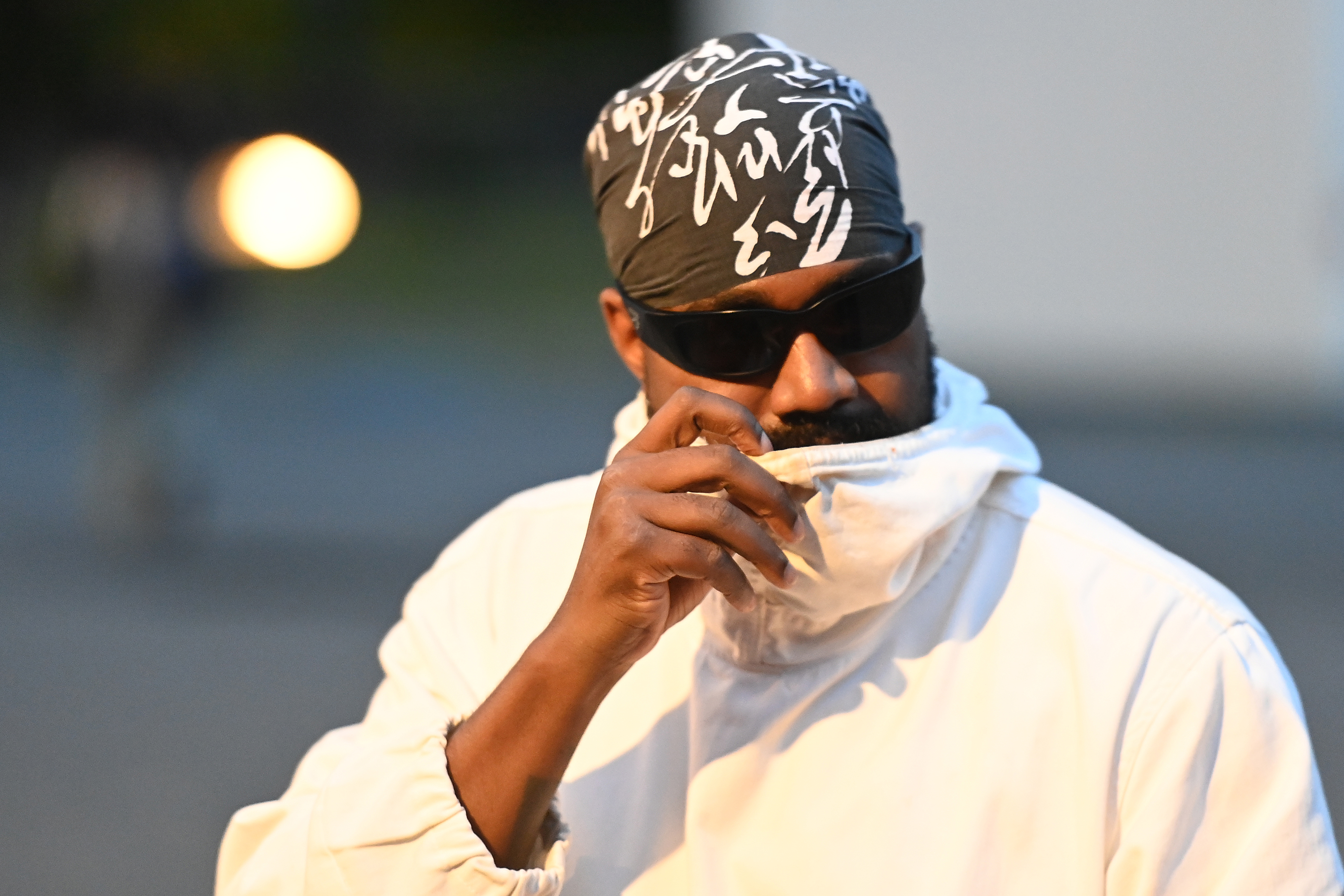 Kanye West wearing sunglasses, a patterned bandana, and a white jacket, partially covering his face with the jacket collar. Kanye West has a history of apologizing for his actions.