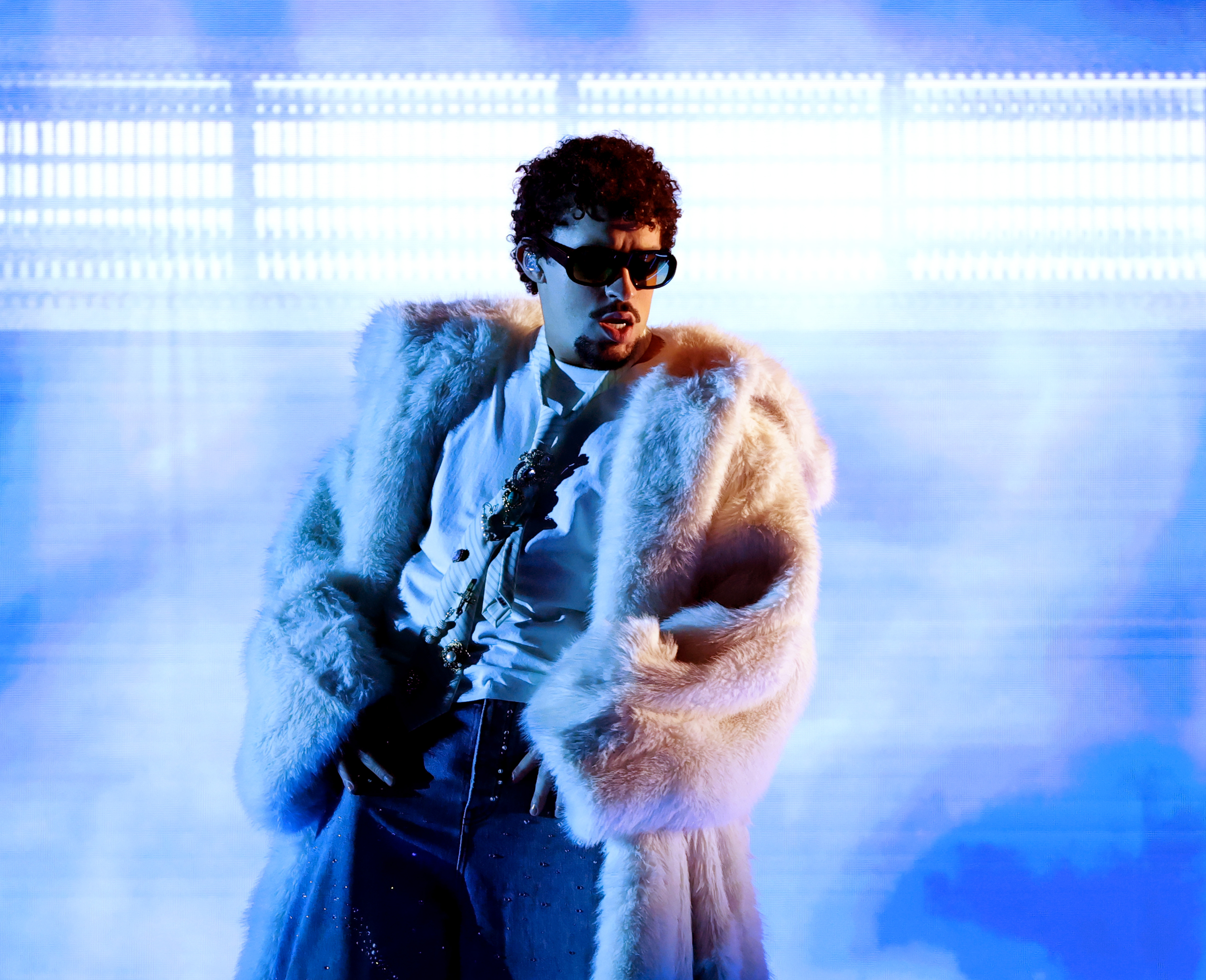 Bad Bunny in a white fur coat and sunglasses performs on stage with blue lighting effects. He is getting ready to play the Super Bowl. He was the most streamed artist on Spotify in 2025, on fact you might not have known about Bad Bunny.