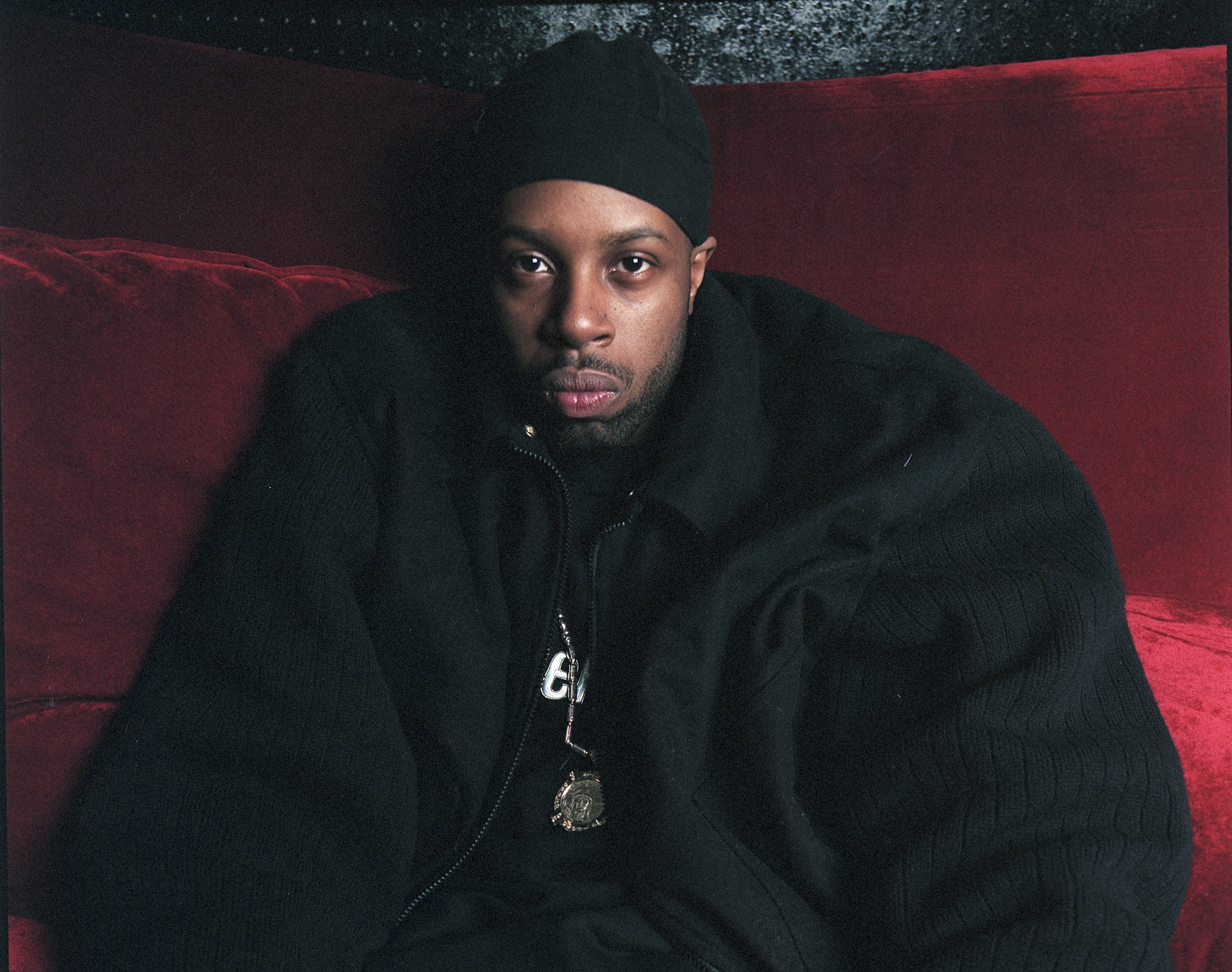 J Dilla wearing a black beanie and jacket sits on a red couch, looking directly at the camera. He is one of the greatest producers of all time. Here is the 50 greatest J Dilla songs.