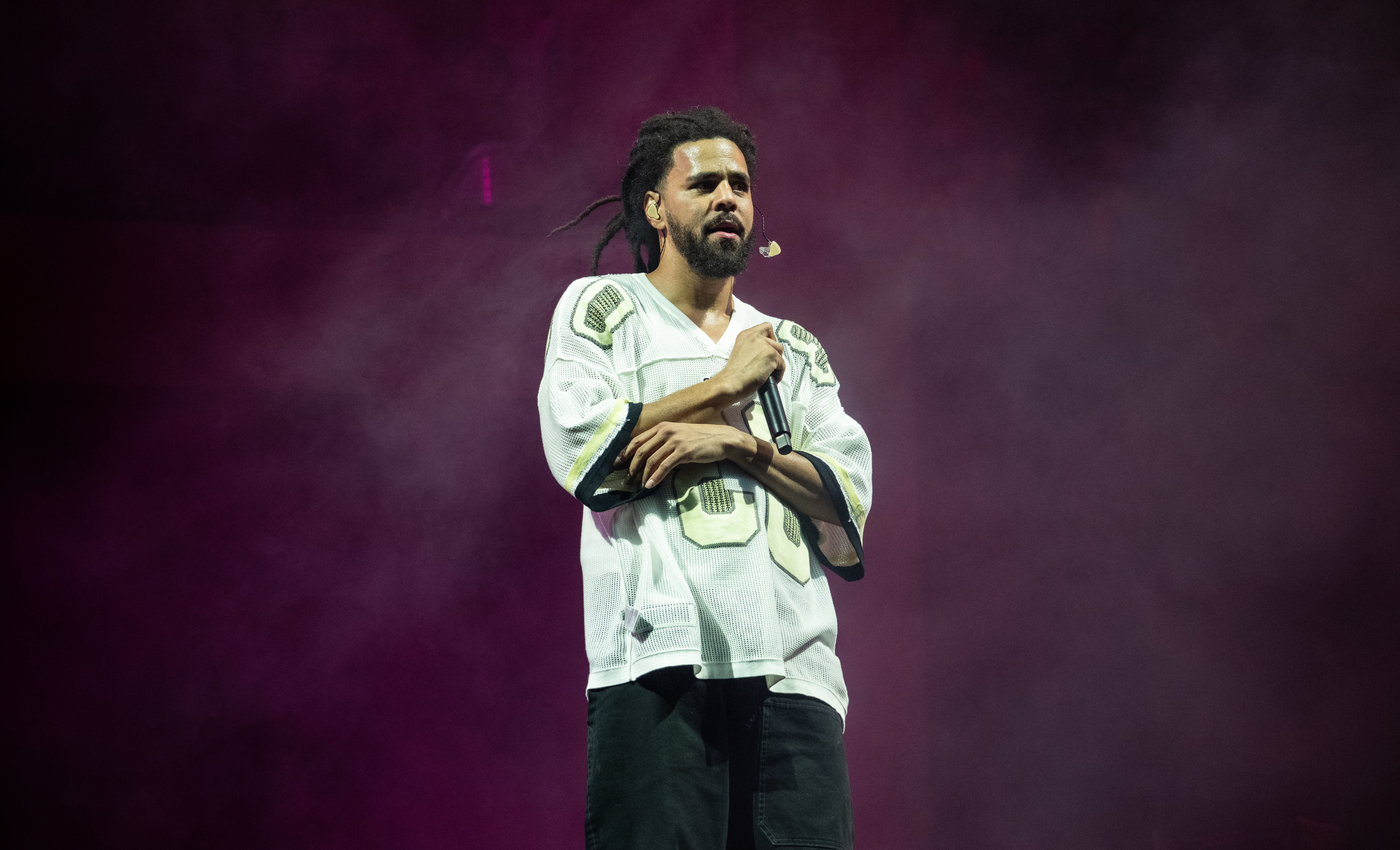 J. Cole, who has released albums like 2014 Forest Hills Drive and The Fall Off, performing on stage, wearing a white jersey and holding a microphone, with a smoky purple background. 