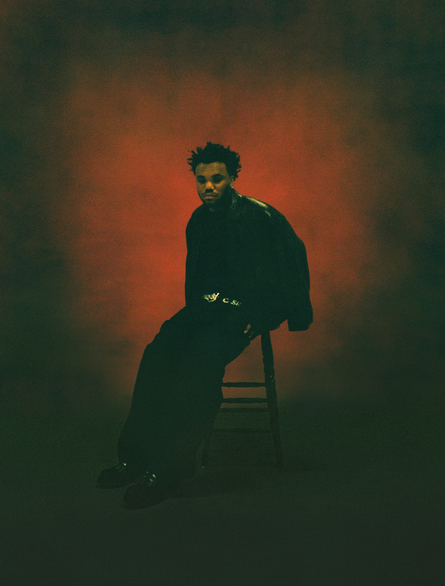 Baby Keem, who just released his new album Ca$ino, is sitting on a chair against a dark, moody red background, wearing a dark outfit with a jacket and belt. He has a sad face on.