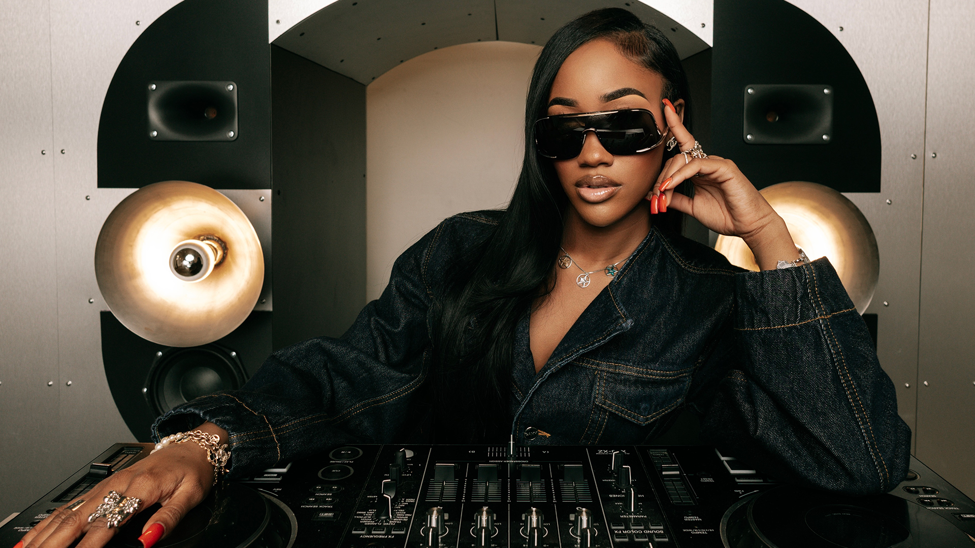 A woman in sunglasses and a denim jacket poses confidently at a DJ booth with large speakers in the background.