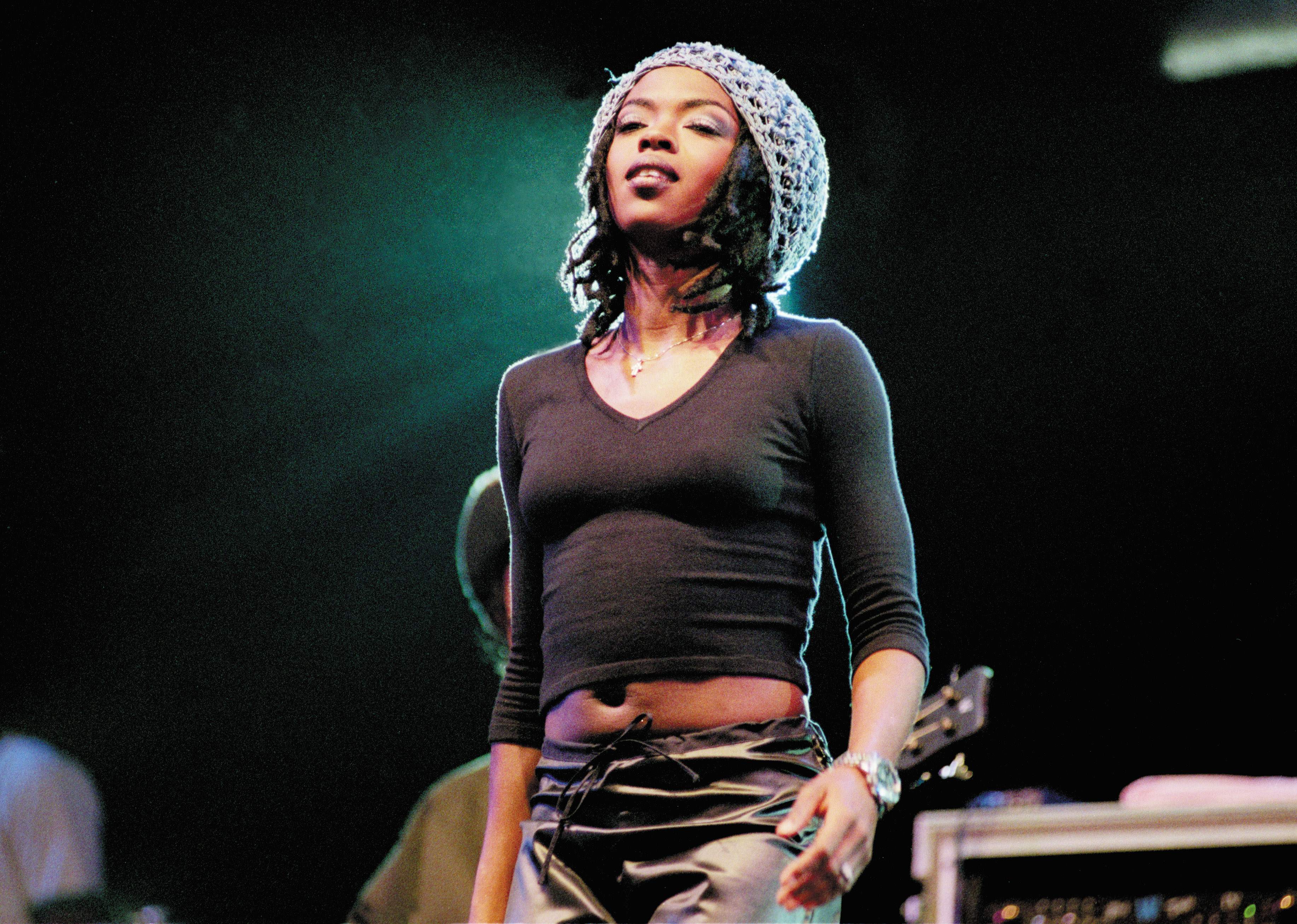 Lauryn Hill performing on stage, wearing a black top and a knit cap, with a confident expression. She was just nominated for the rock and roll hall of fame.