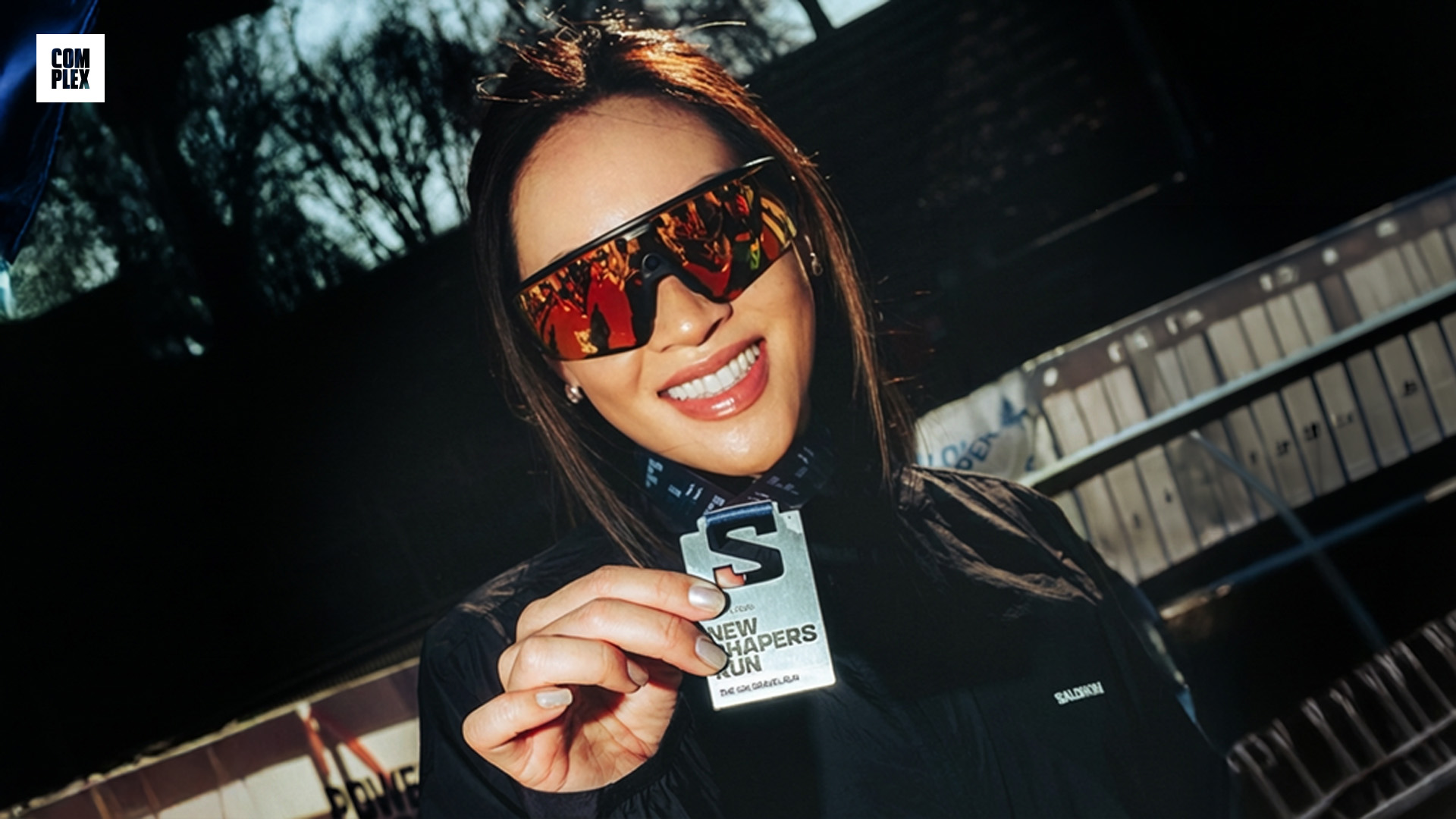 A woman wearing reflective sunglasses holds up a medal with "New Runners Run" on it, smiling outdoors.