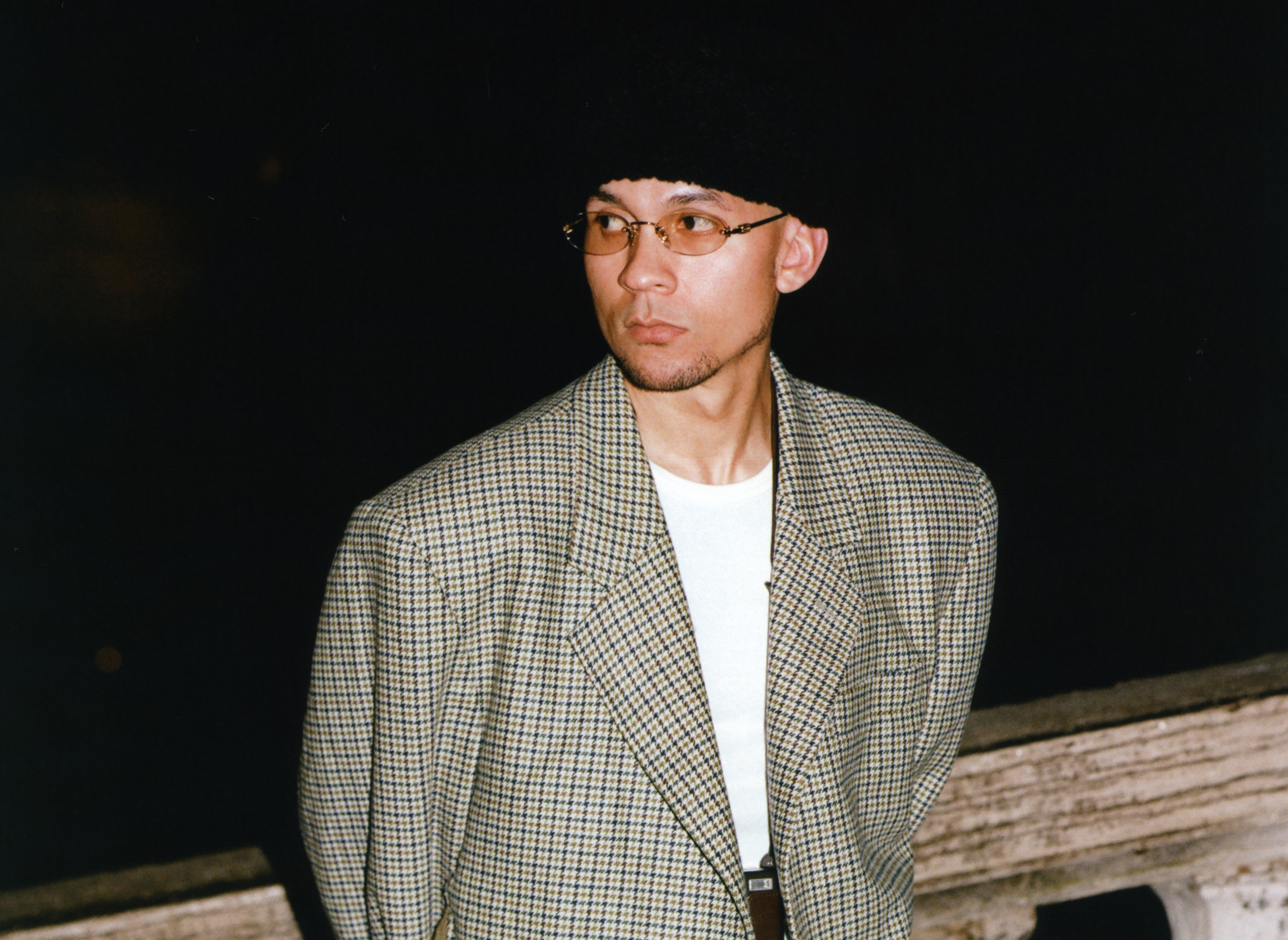 Tainy is wearing a checkered blazer, glasses, and a black hat, looking to the side against a dark background. He has produced some of the most iconic reggaeton songs of all time.