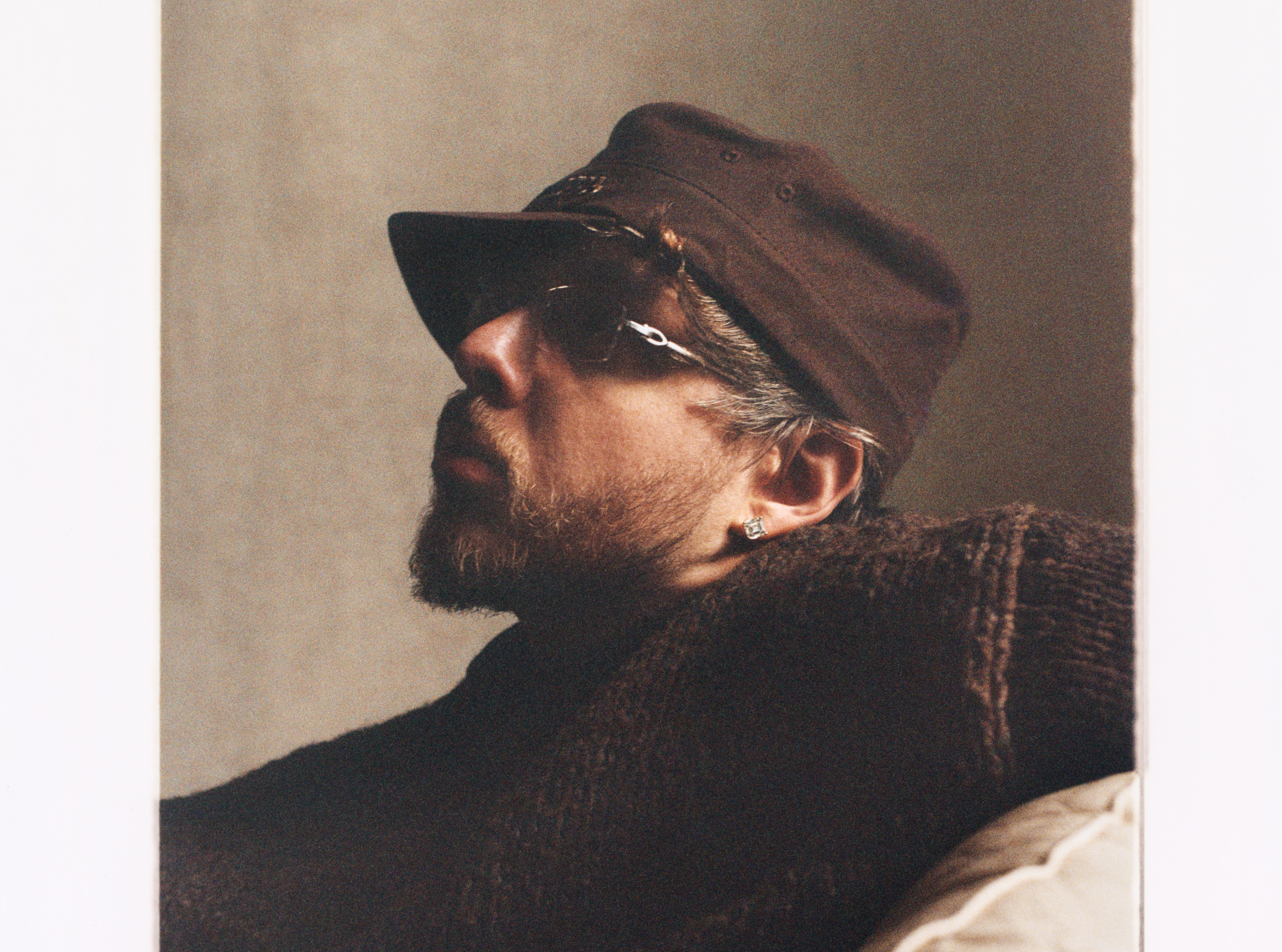 Jack Harlow wearing a brown cap and sunglasses, with a beard and earring, reclining on a couch, in a relaxed pose. These are photos from his new album Monica.