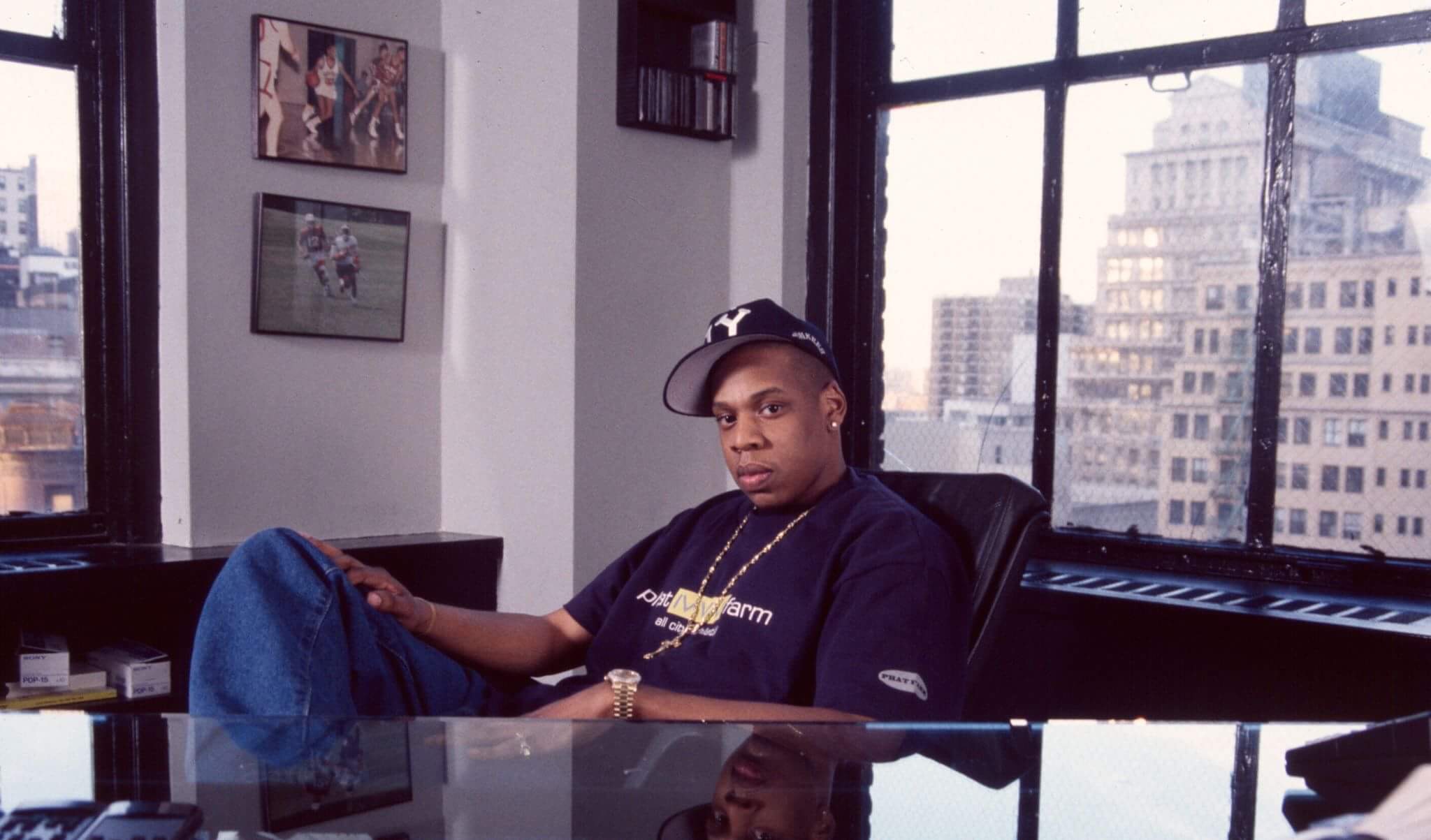Jay-Z sitting in an office, wearing a cap and casual attire, with cityscape visible through large windows behind him. This was during the Reasonable Doubt era, which is one of the greatest rap albums of the '90s.