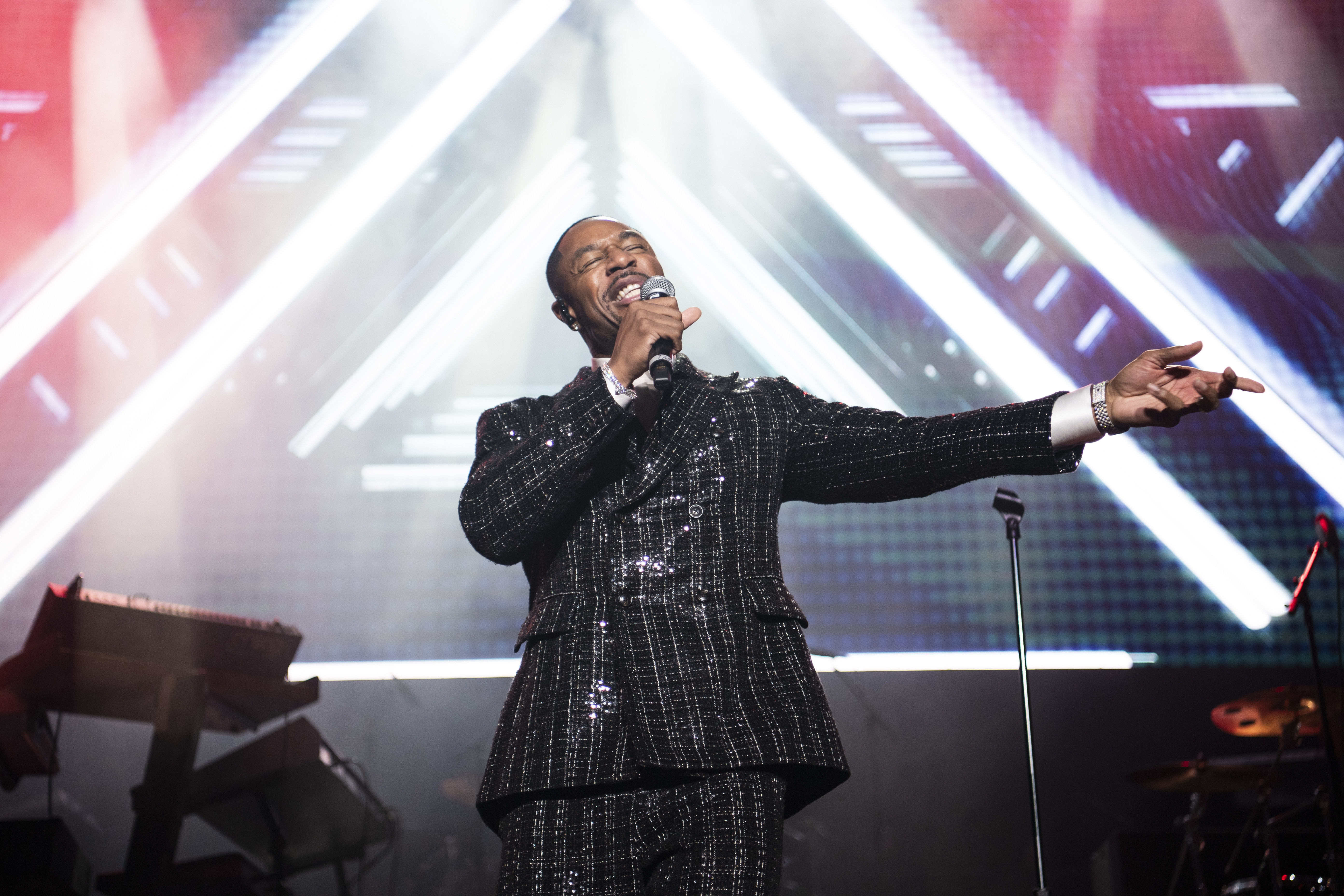 Tank in a sparkling suit sings passionately on stage with dynamic lighting and geometric patterns in the background. He is getting ready to face off against Tyrese for VERZUZ.