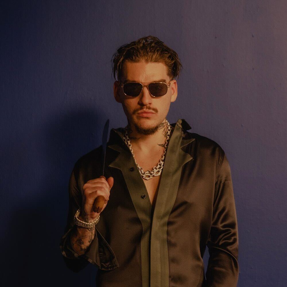 Rapper Yeat wearing sunglasses and a dark satin shirt holding a knife, posing against a blue background. This is a photo attached to his latest album, ADL, which is a double album. 