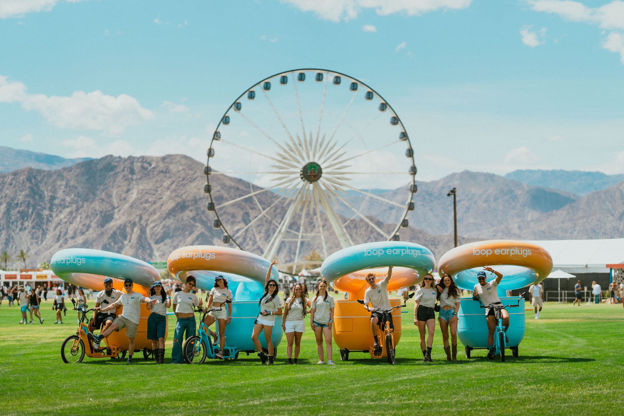 A group of people with pedicabs featuring Loop earplugs branding, in front of a Ferris wheel at a festival, with mountains in the background. This is from Coachella