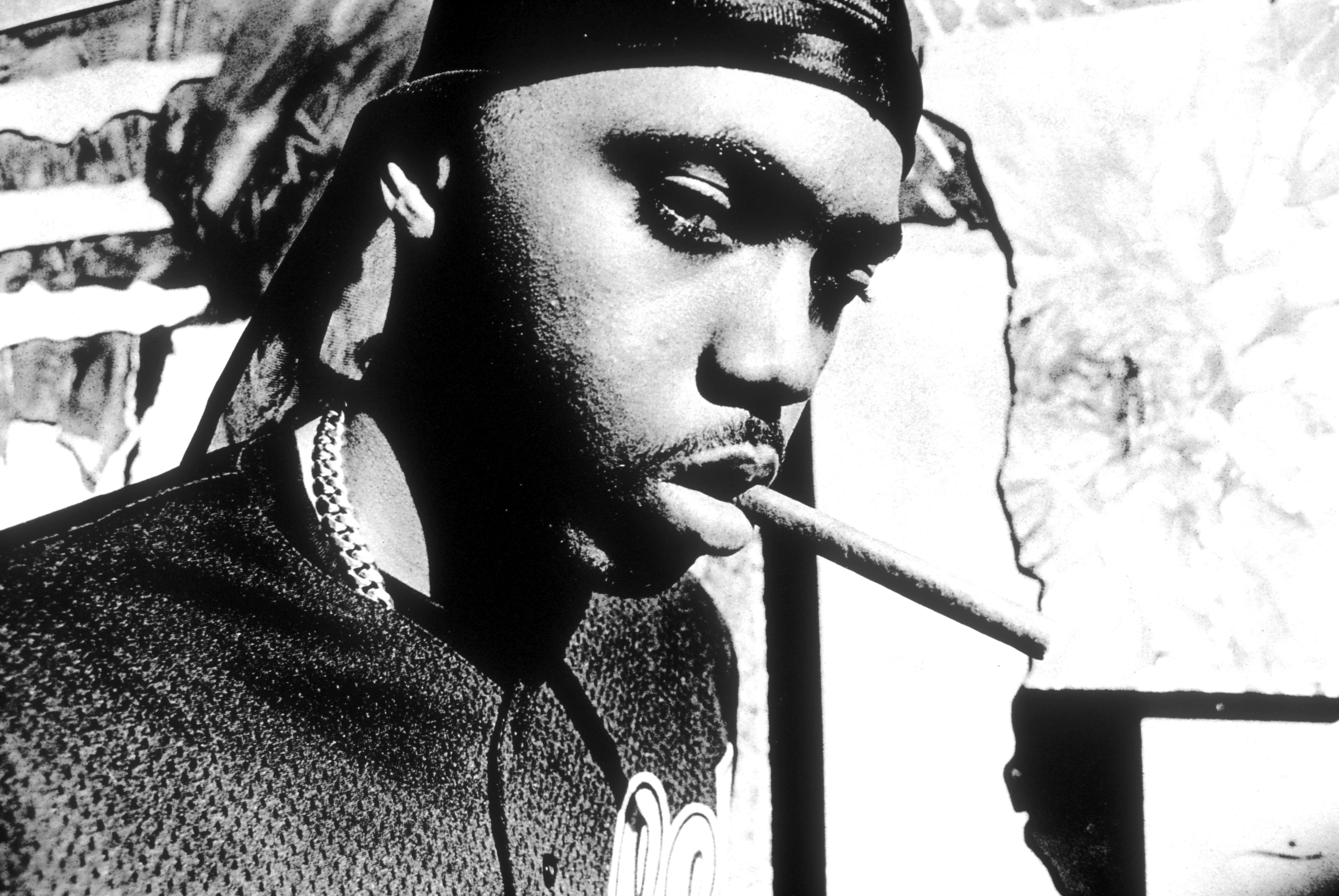 A black and white image of Nas wearing a durag and holding a cigar in his mouth, with a serious expression. This is from Illmatic days, which is Nas' classic debut album.