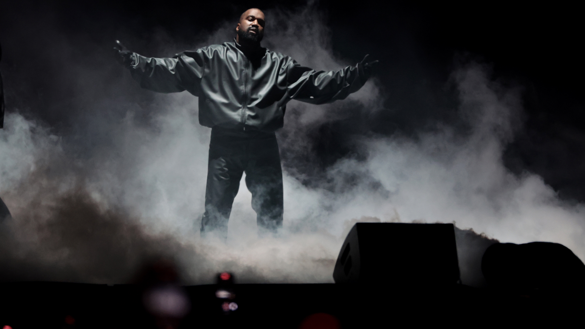 Ye stands on stage with arms outstretched, surrounded by smoke, wearing a dark jacket and pants.
