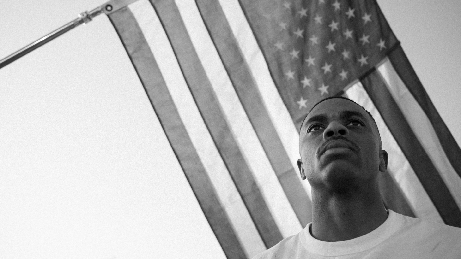 Vince Staples in a white shirt stands in front of a large American flag, looking upward with a serious expression. Black and white image.