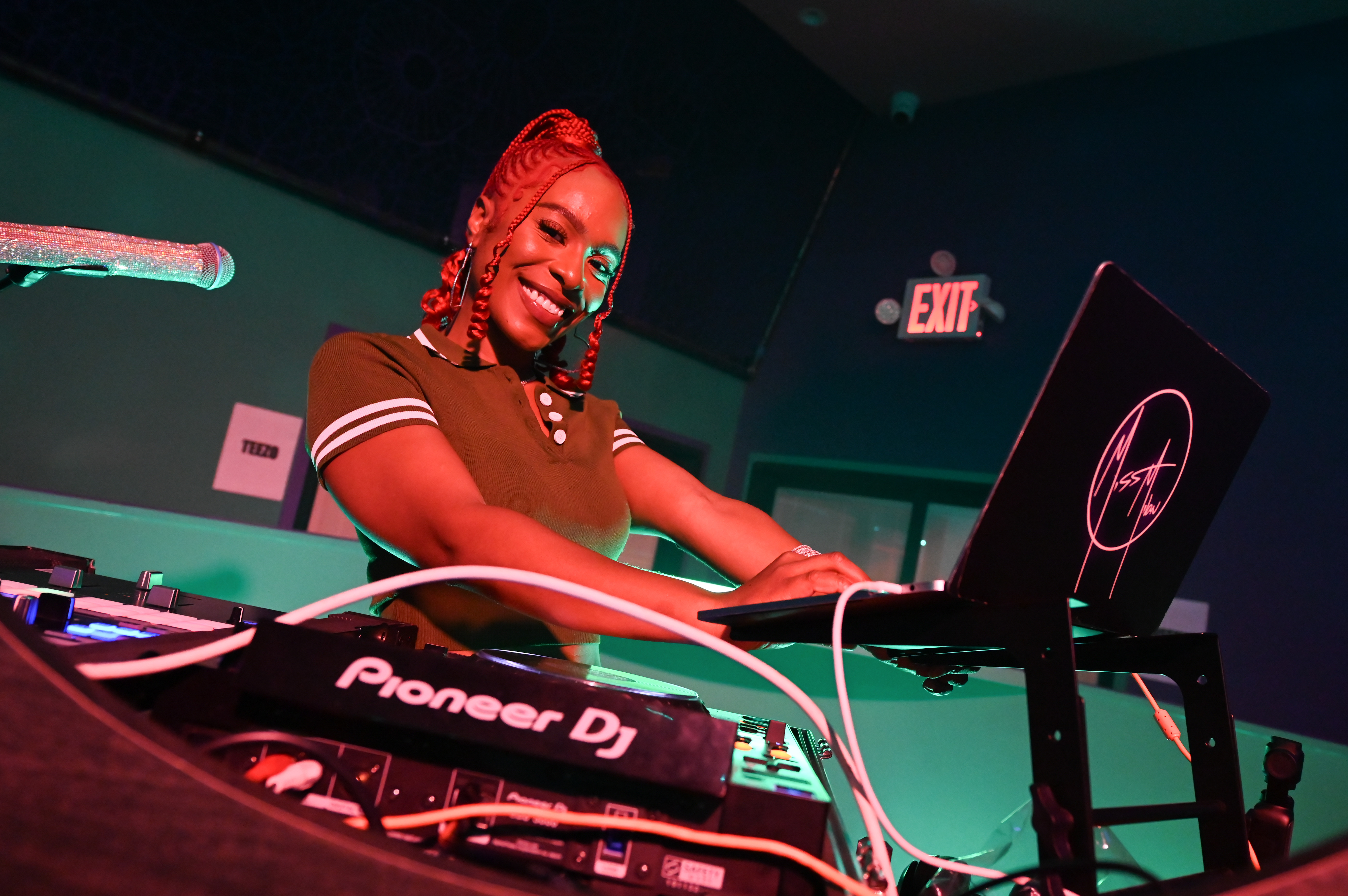 A DJ with braided hair smiles while standing at a DJ booth with Pioneer equipment and a laptop.