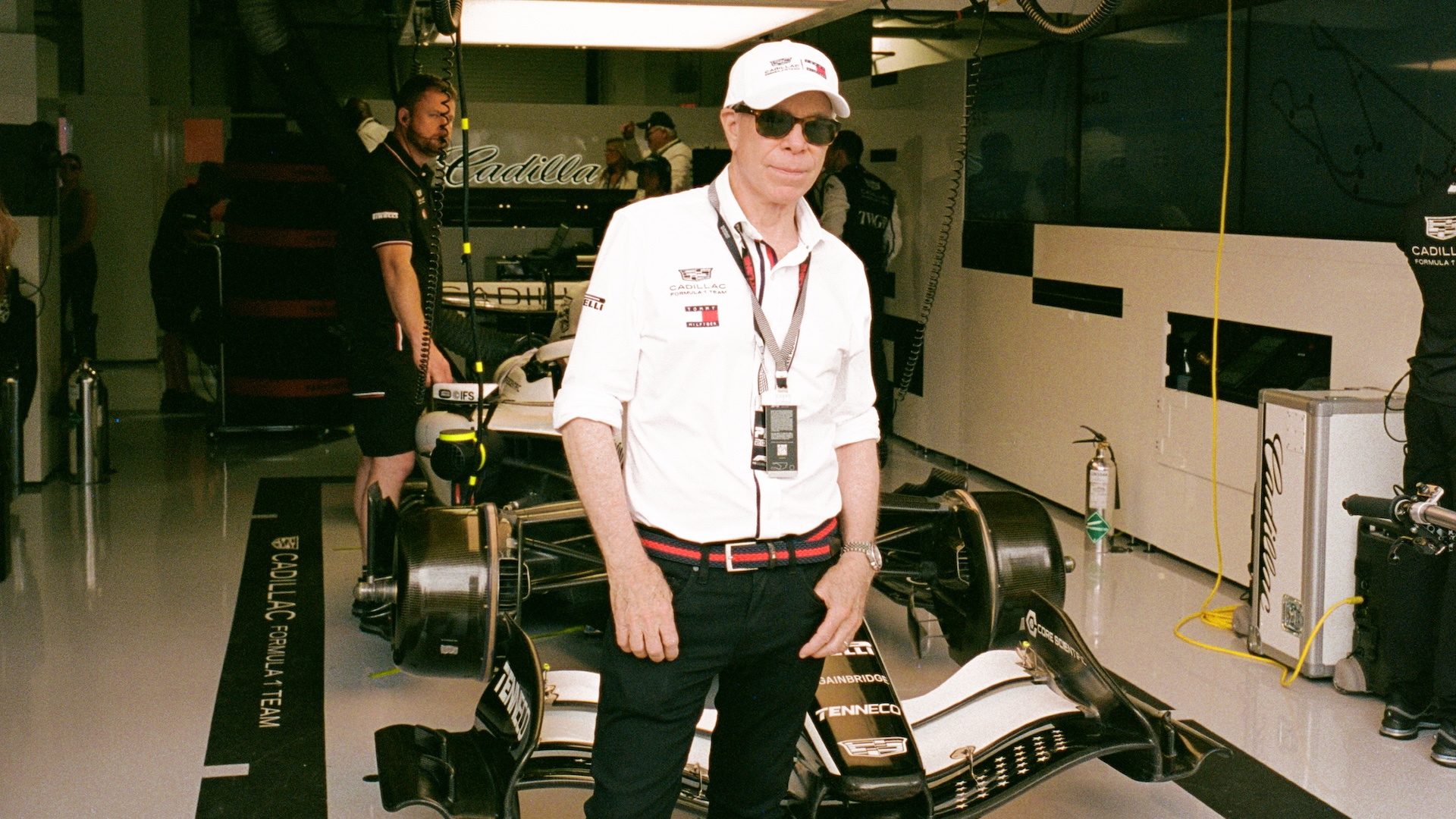 Tommy Hilfiger in a white shirt and cap stands in a race car garage, with a Cadillac race car and team members in the background.