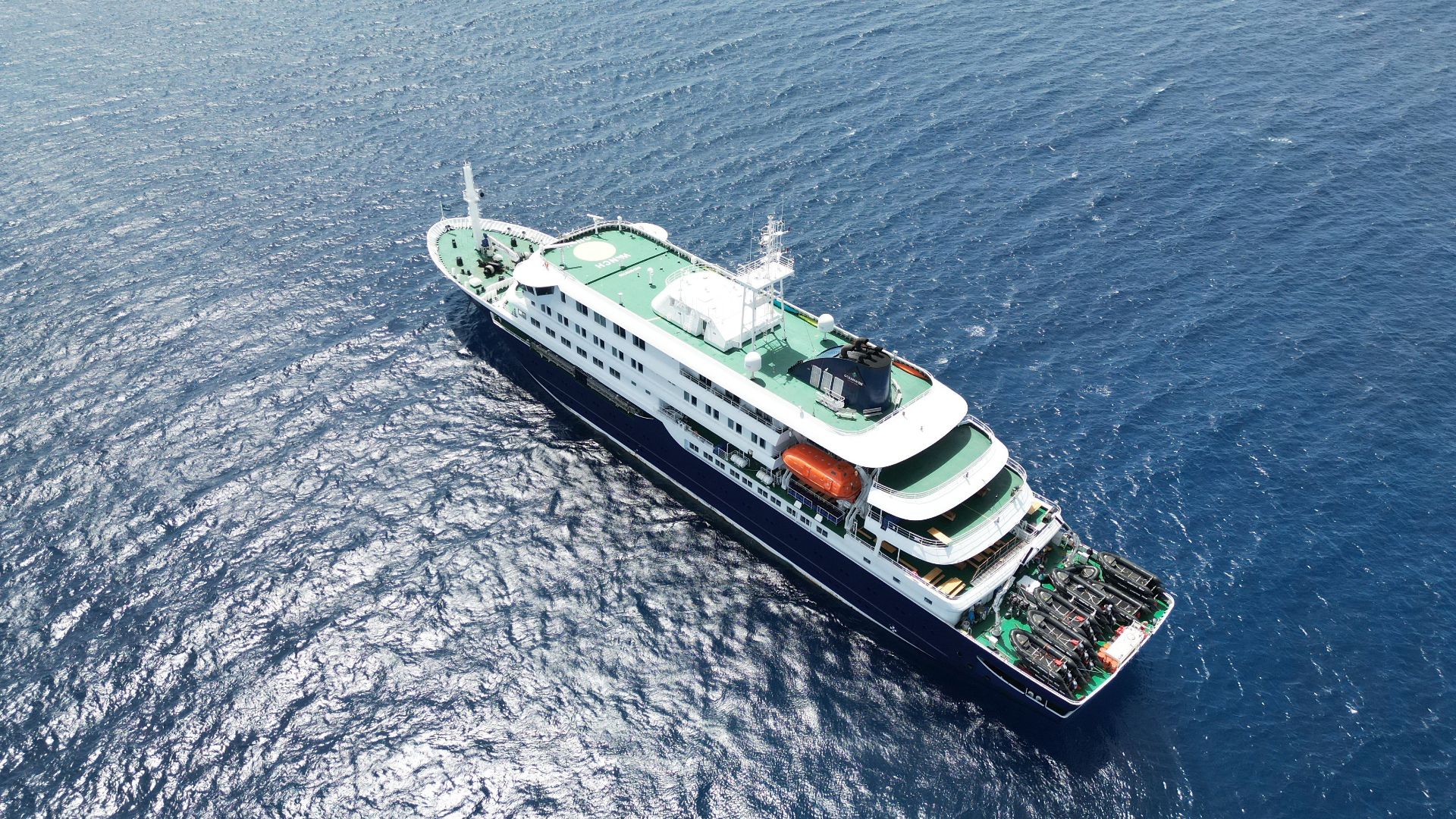 Aerial view of a large cruise ship with a green deck sailing on calm blue ocean waters.