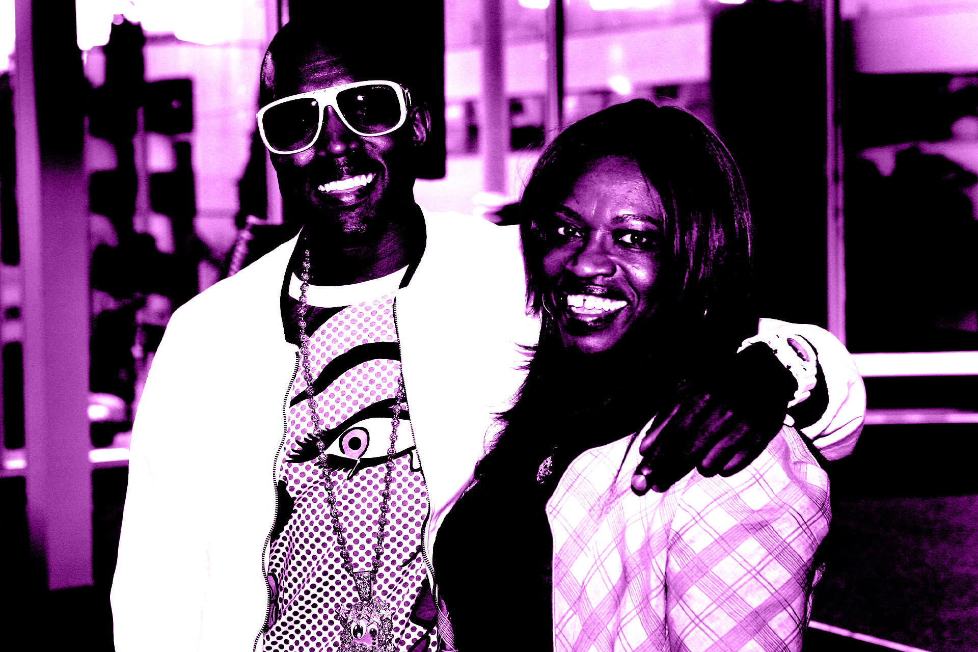 Kanye West and his mom Donda smiling, with the Kanye wearing sunglasses and a patterned shirt. The image has a purple tint.