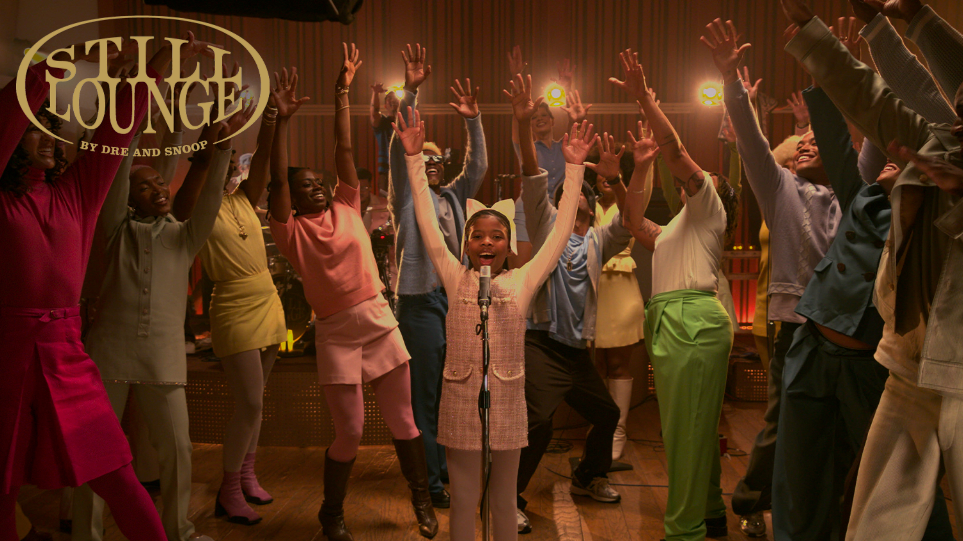 A group of people in colorful outfits are joyfully raising their hands on a stage. A child is singing into a microphone.
