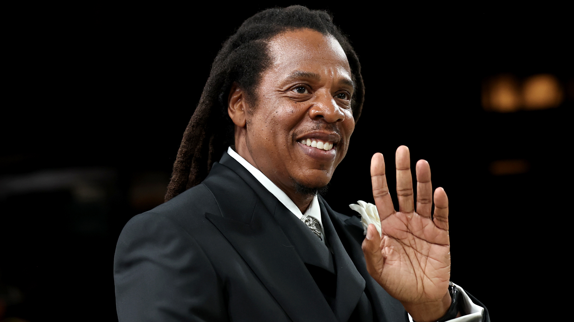 Jay-Z smiling and waving, wearing a black suit with a white shirt and tie, against a dark background.
