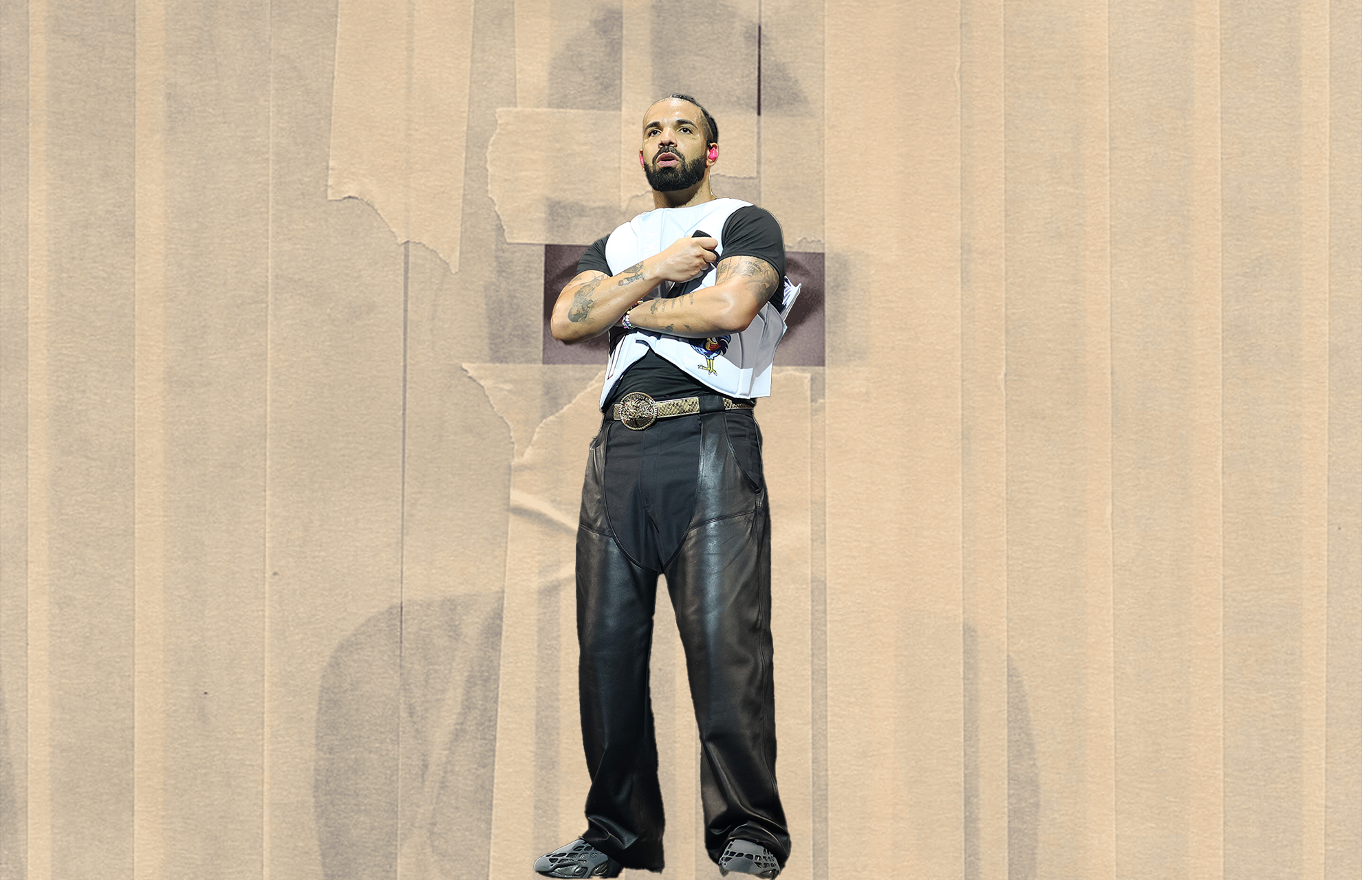 Drake with tattoos stands against a textured backdrop, wearing a black and white shirt, leather pants, and a belt with a prominent buckle.