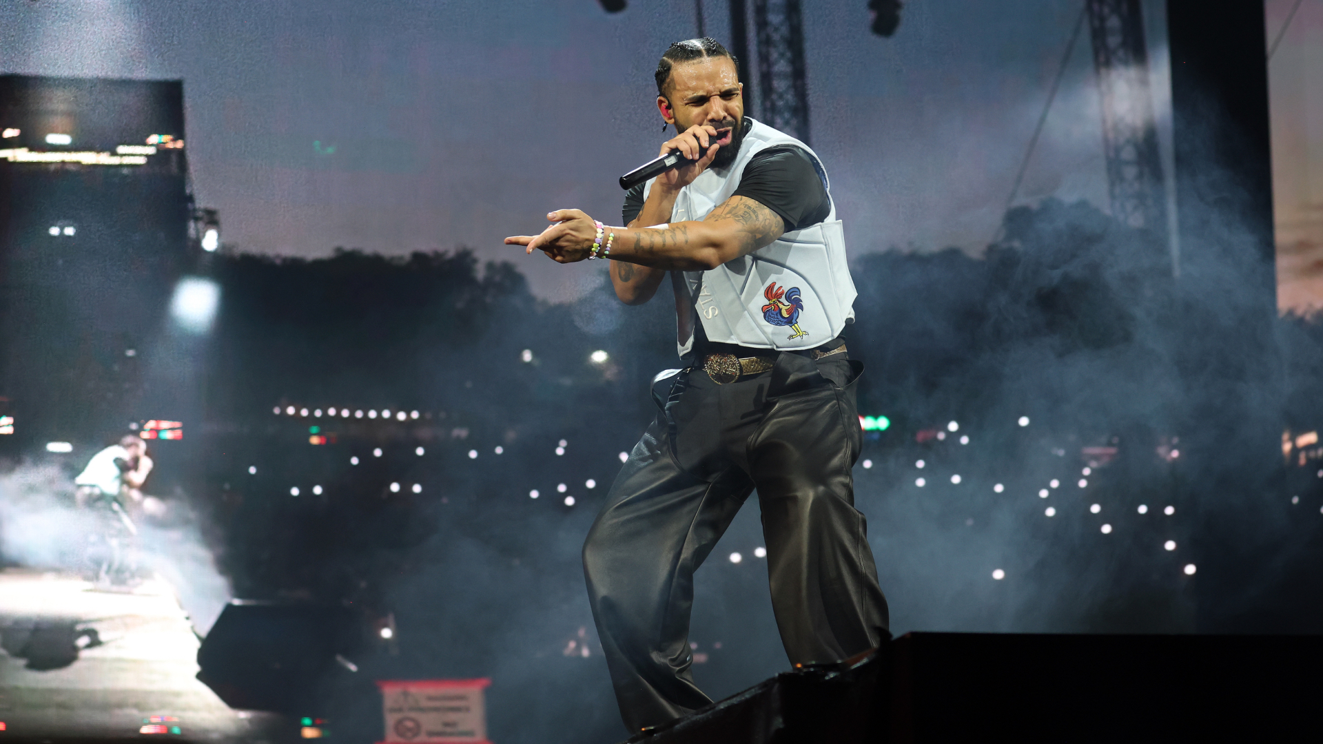 Drake with cornrows and tattoos sings energetically on stage, wearing a white vest and black pants. The crowd is visible in the background.
