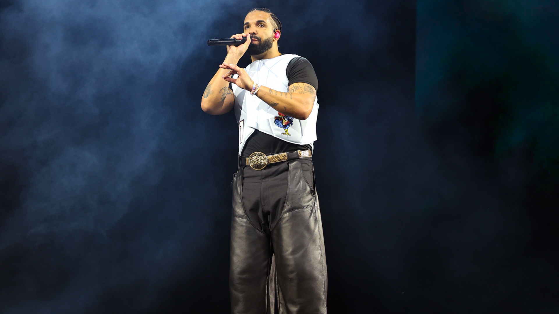 Drake performing on stage, wearing a white vest over a black shirt and leather pants, holding a microphone.