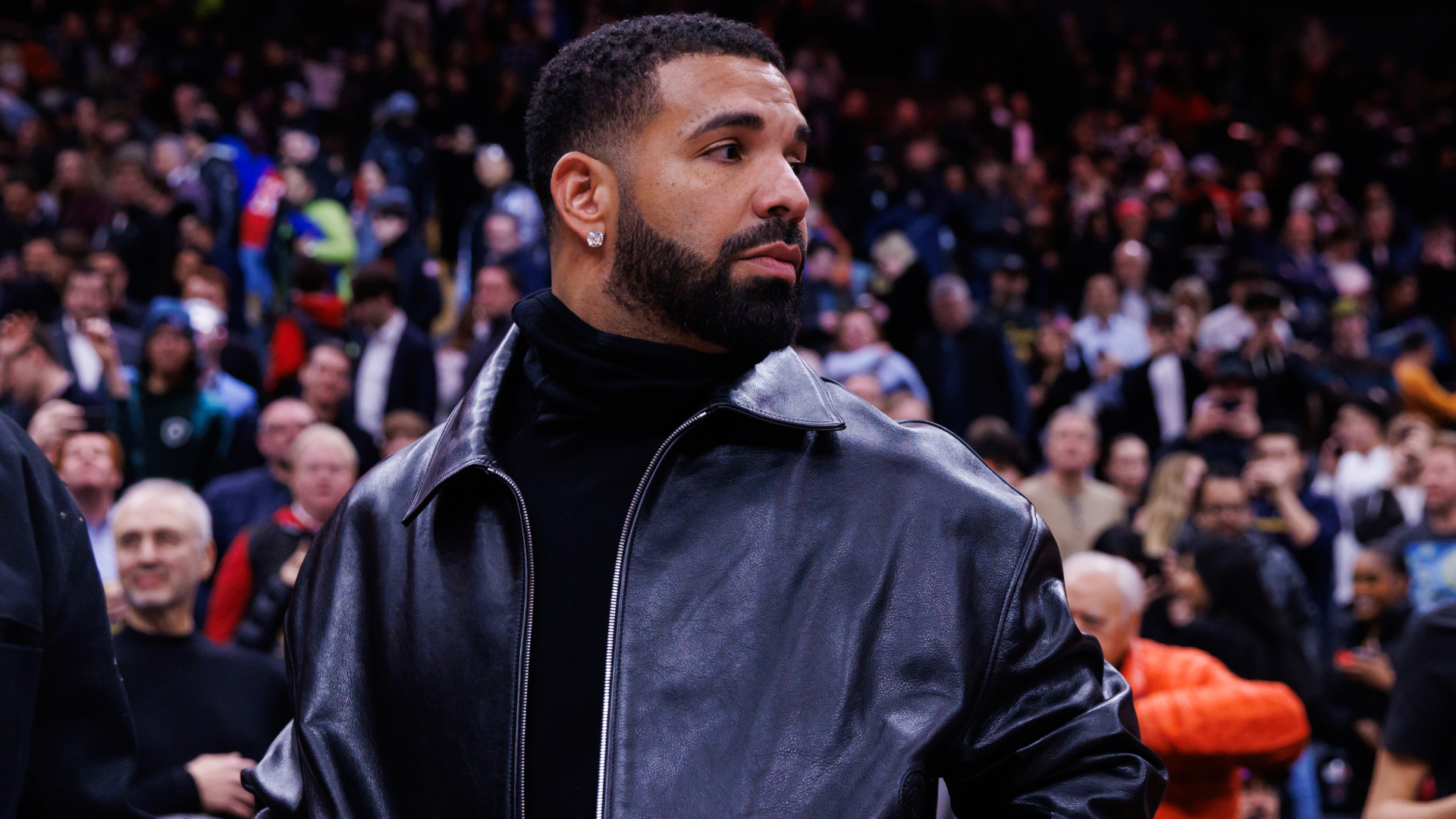 Drake wearing a black leather jacket, standing in a crowded venue with people in the background.