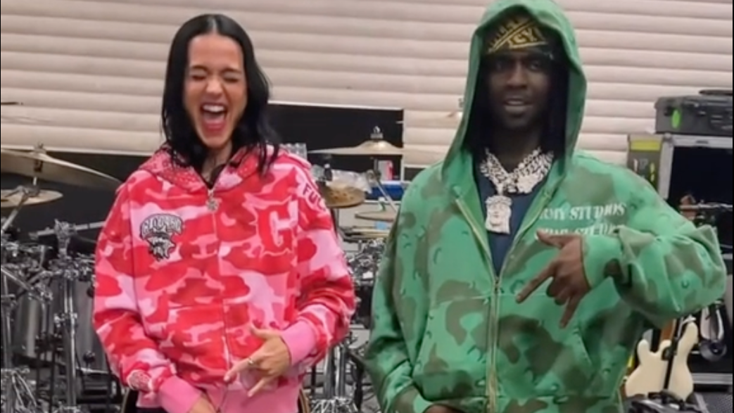 Two people in colorful hoodies stand in a music studio. The woman in pink is laughing, and the man in green poses with hand gestures.