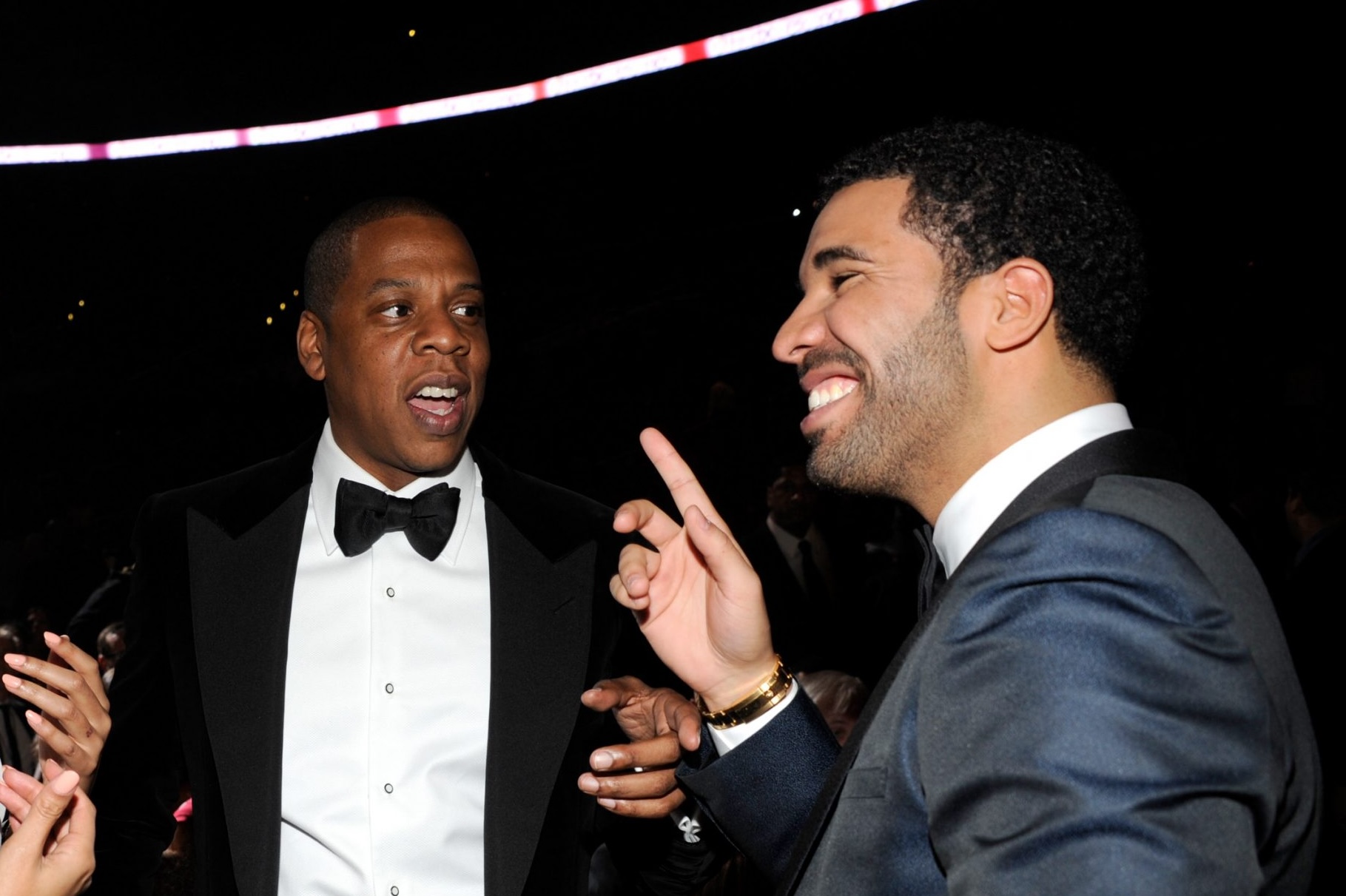Jay-Z in a tuxedo talking to Drake, who is smiling and gesturing, at a formal event.