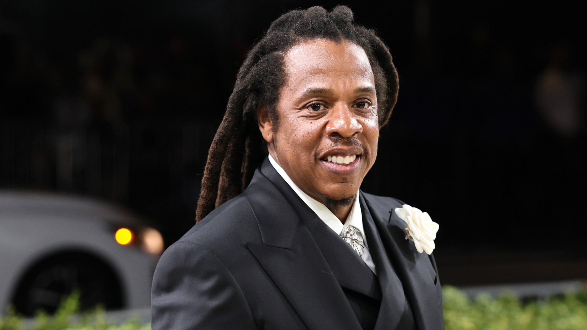 Jay-Z with long dreadlocks in a formal black suit, smiling, with a white flower on his lapel.
