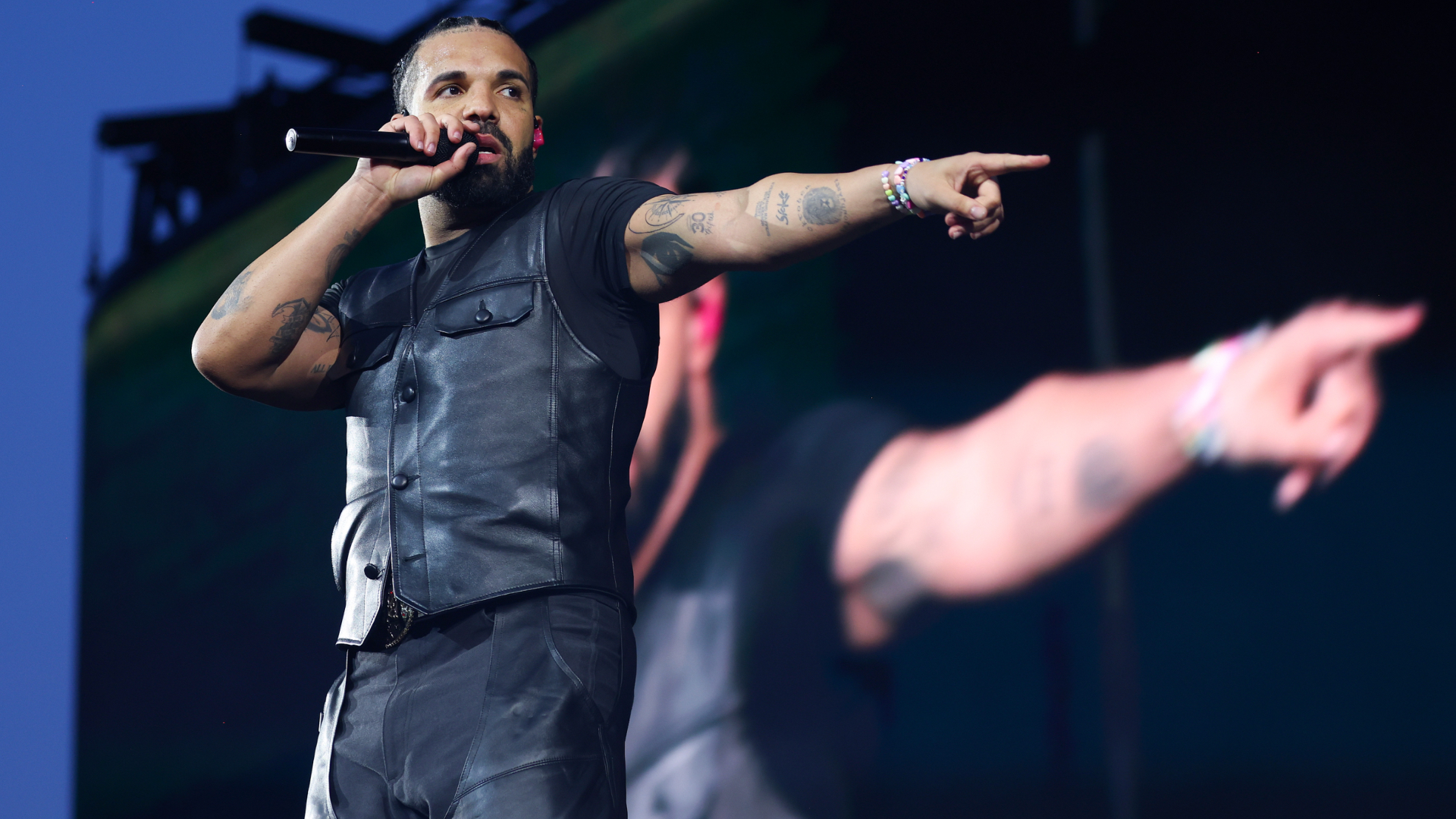 Drake wearing a leather vest and holding a microphone points to the crowd on stage, with a large screen behind them.