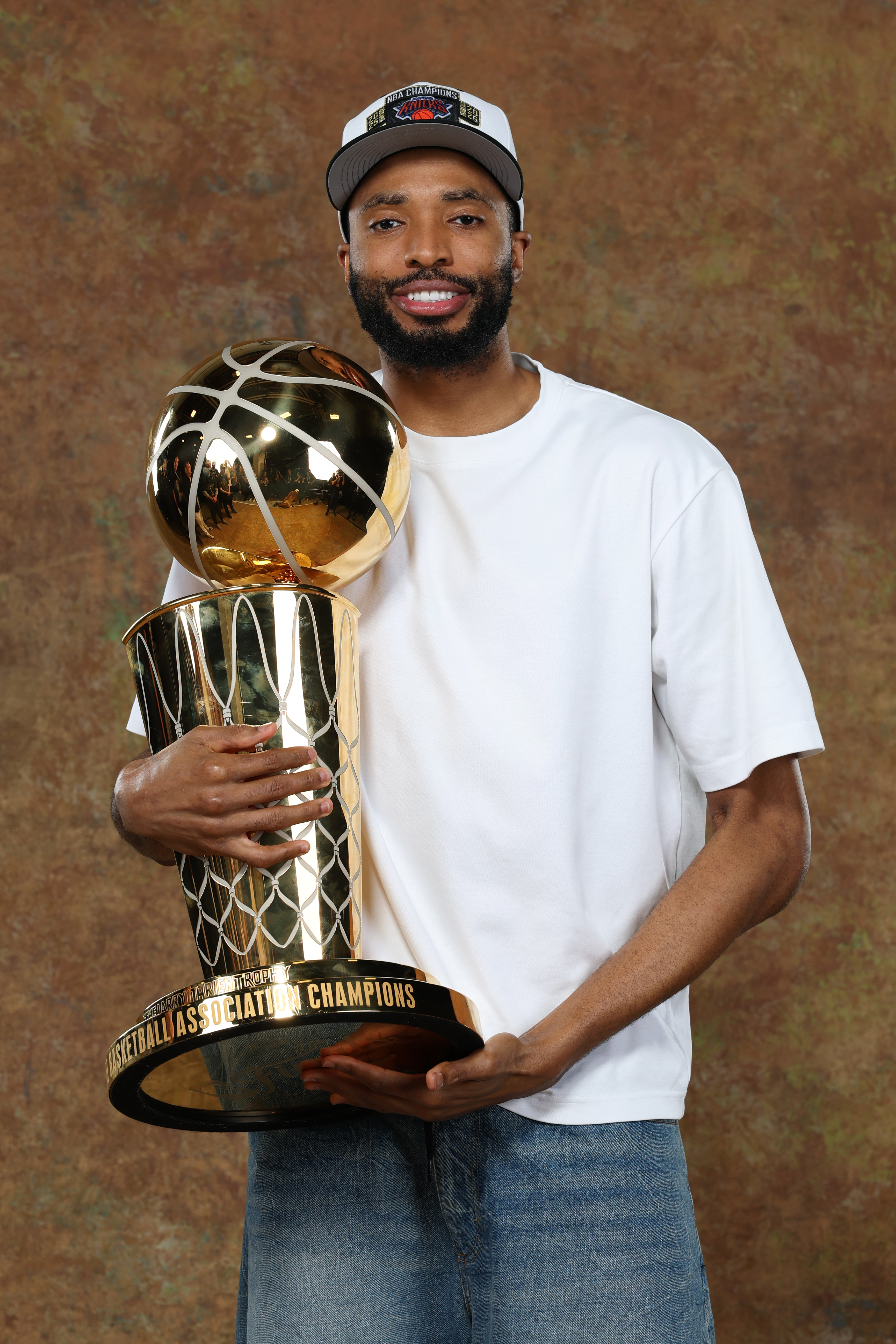 Mikal Bridges, in a white shirt and cap, holds a large basketball trophy, smiling.