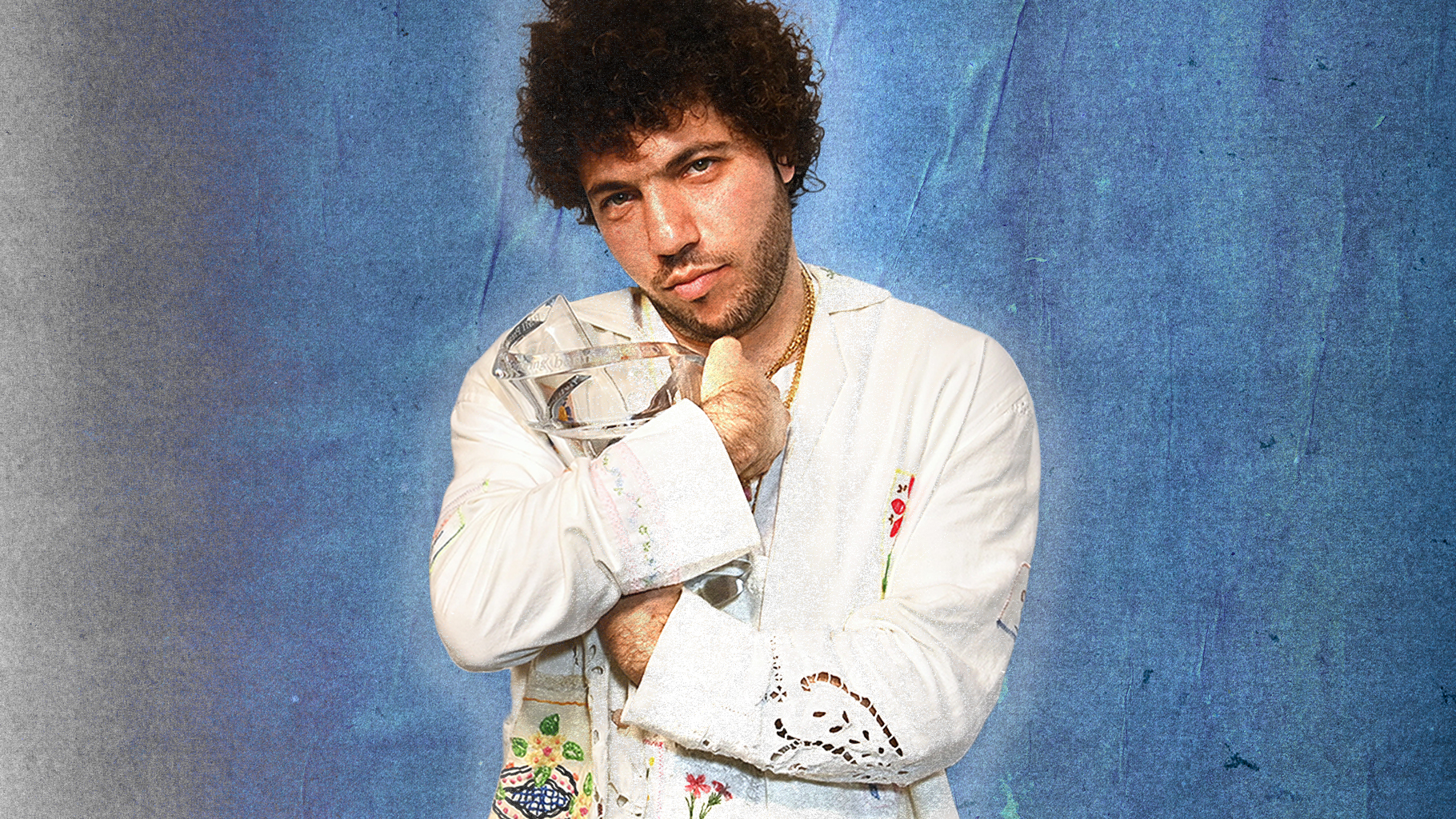 Benny Blanco, who has curly hair, in a white embroidered shirt holds a crystal object against a blue textured background.