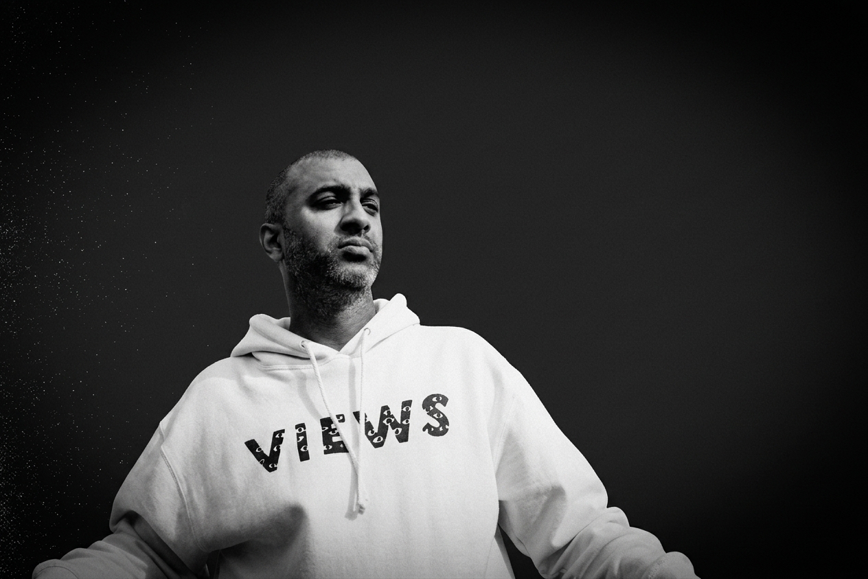 Drake producer Maneesh in a "VIEWS" hoodie stands against a dark background, looking contemplative. Black and white image. He was a producer on the Drake album 'Iceman.'